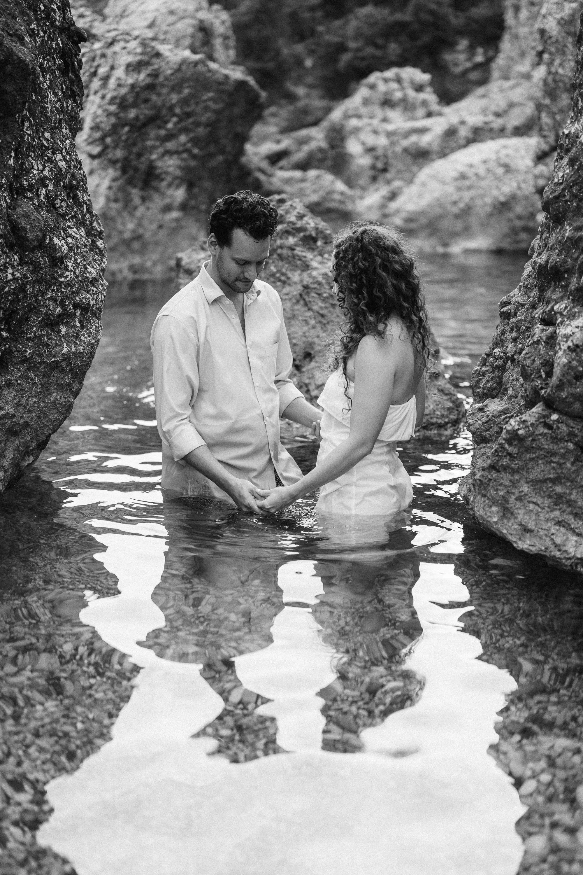 A black and white photo of a couple standing in a shallow river, holding hands, surrounded by rocks with reflections of their figures in the water.