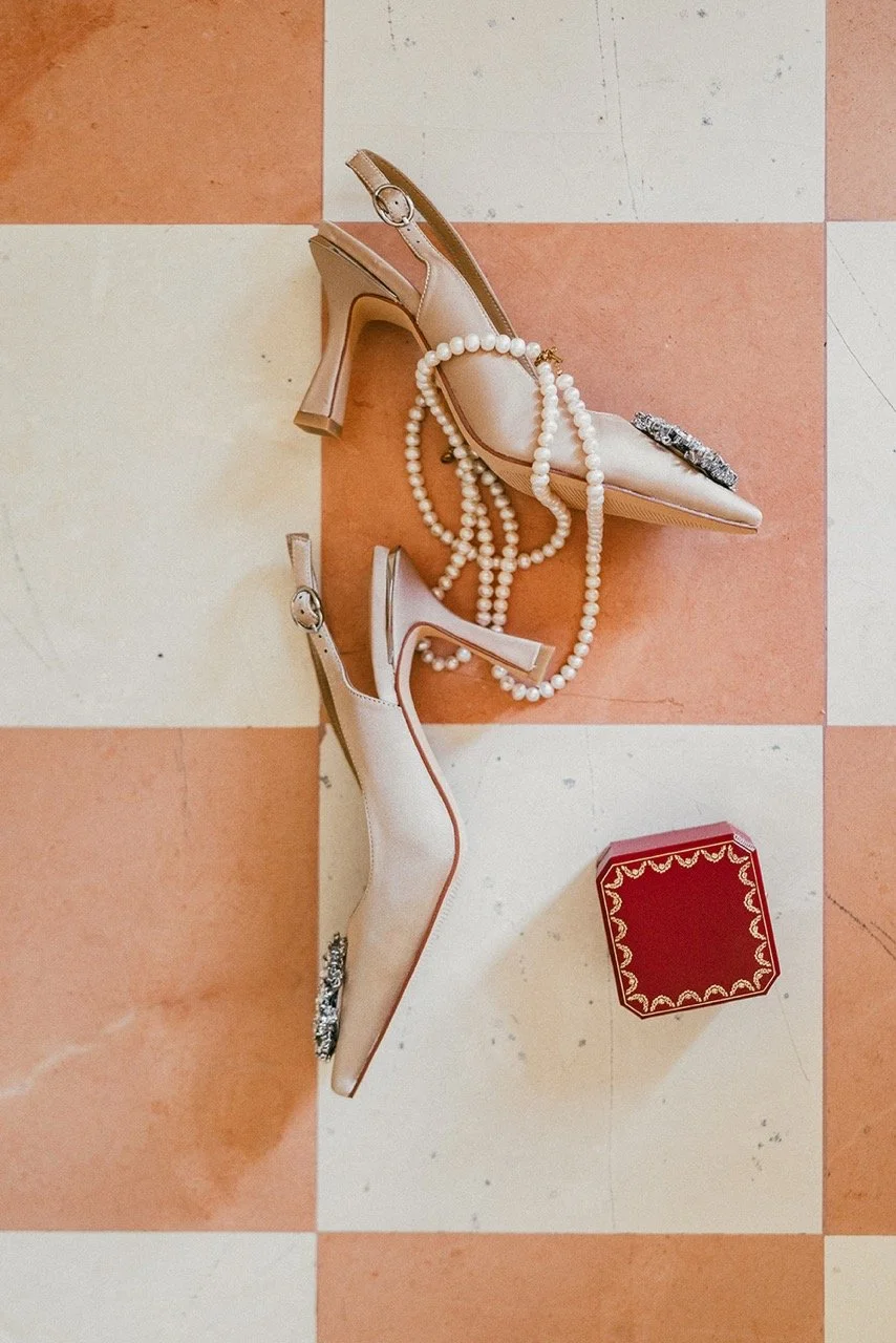 Pair of beige high-heeled shoes with pearl necklaces and jewelry laid out on a checkered tile floor, with a small red box nearby.