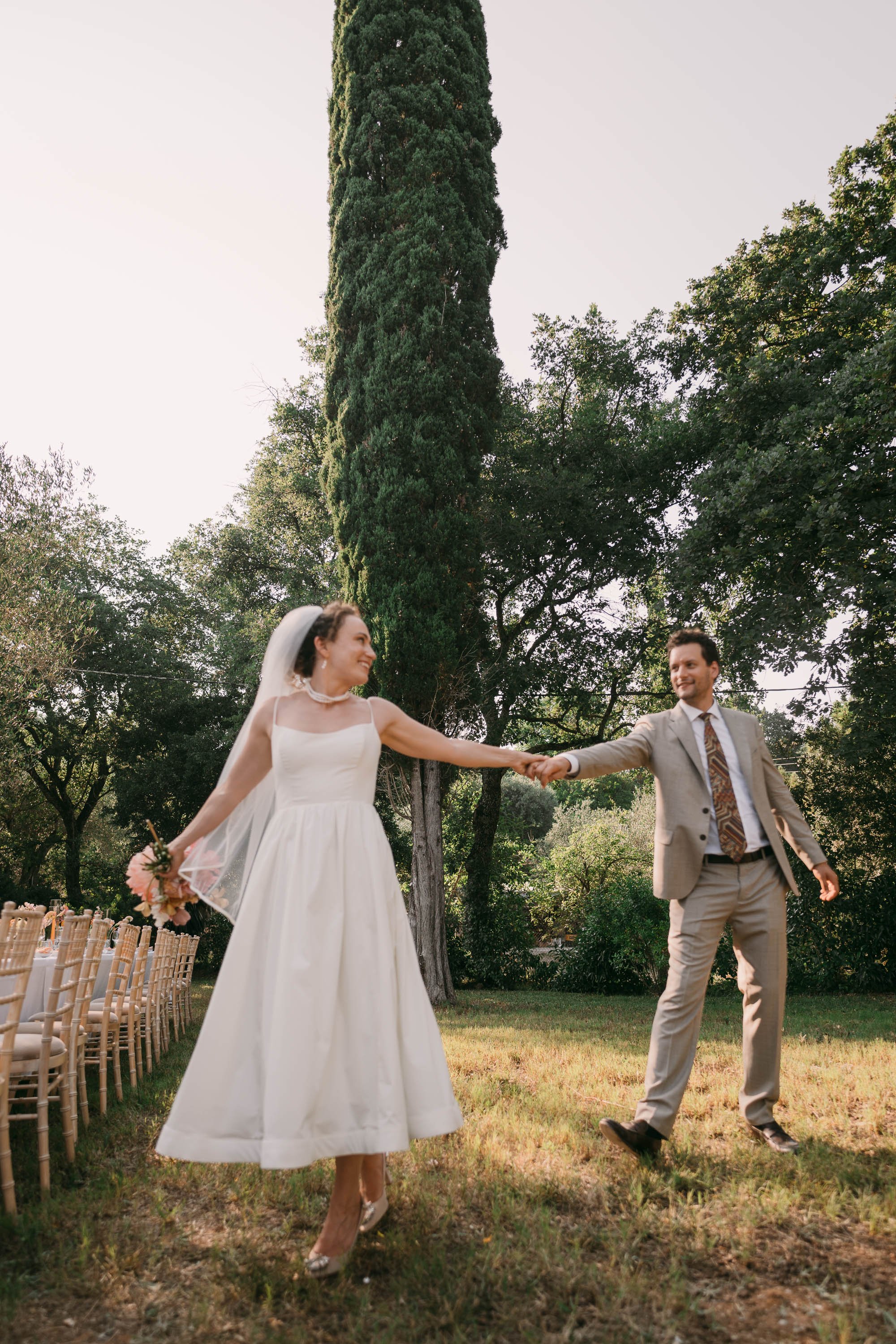 A bride and groom joyfully dancing outdoors on their wedding day, holding hands, with chairs and large trees in the background.