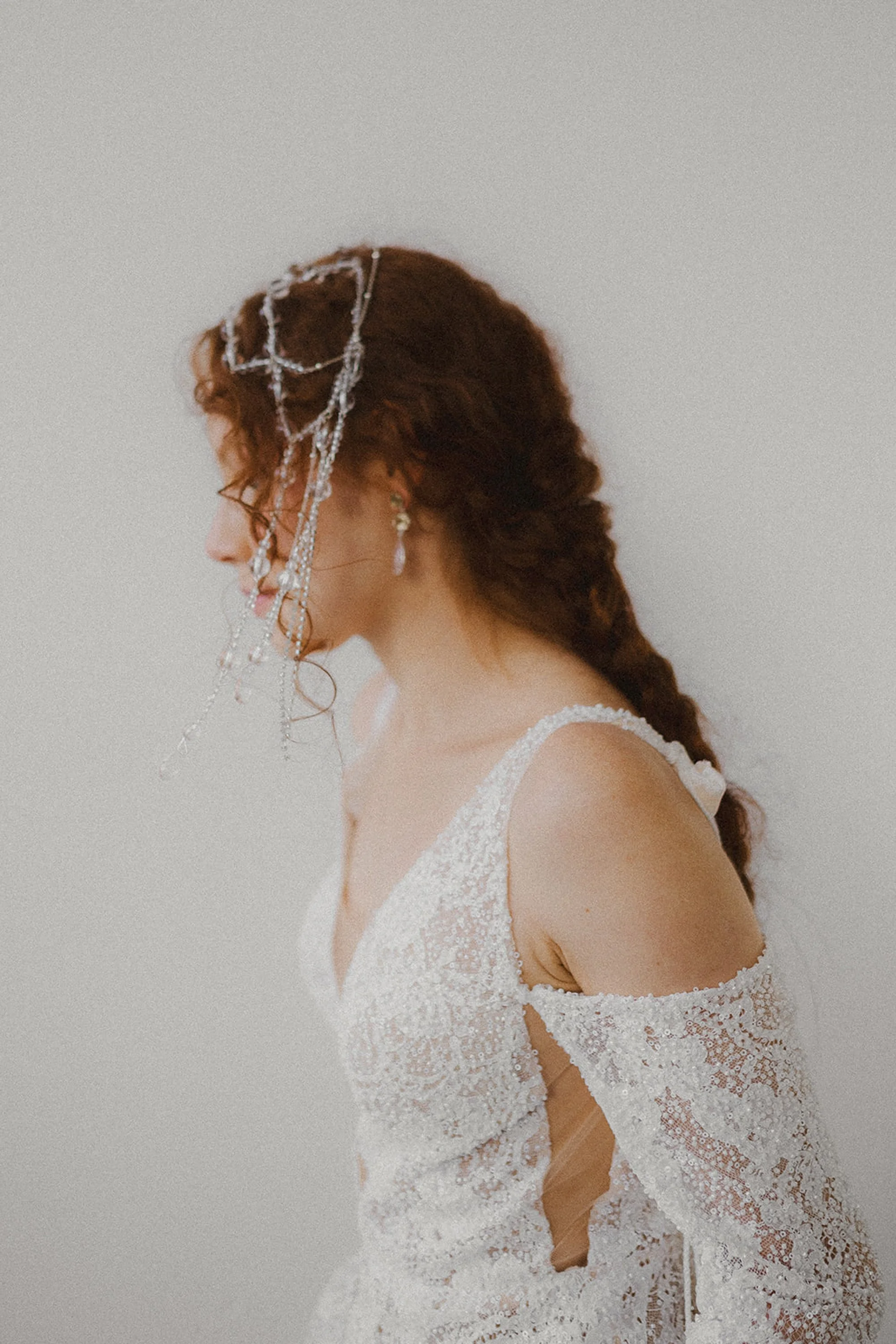 A woman in an elegant lace wedding dress with off-the-shoulder sleeves and intricate embroidery, wearing jewelry and a decorative face chain, against a plain wall.