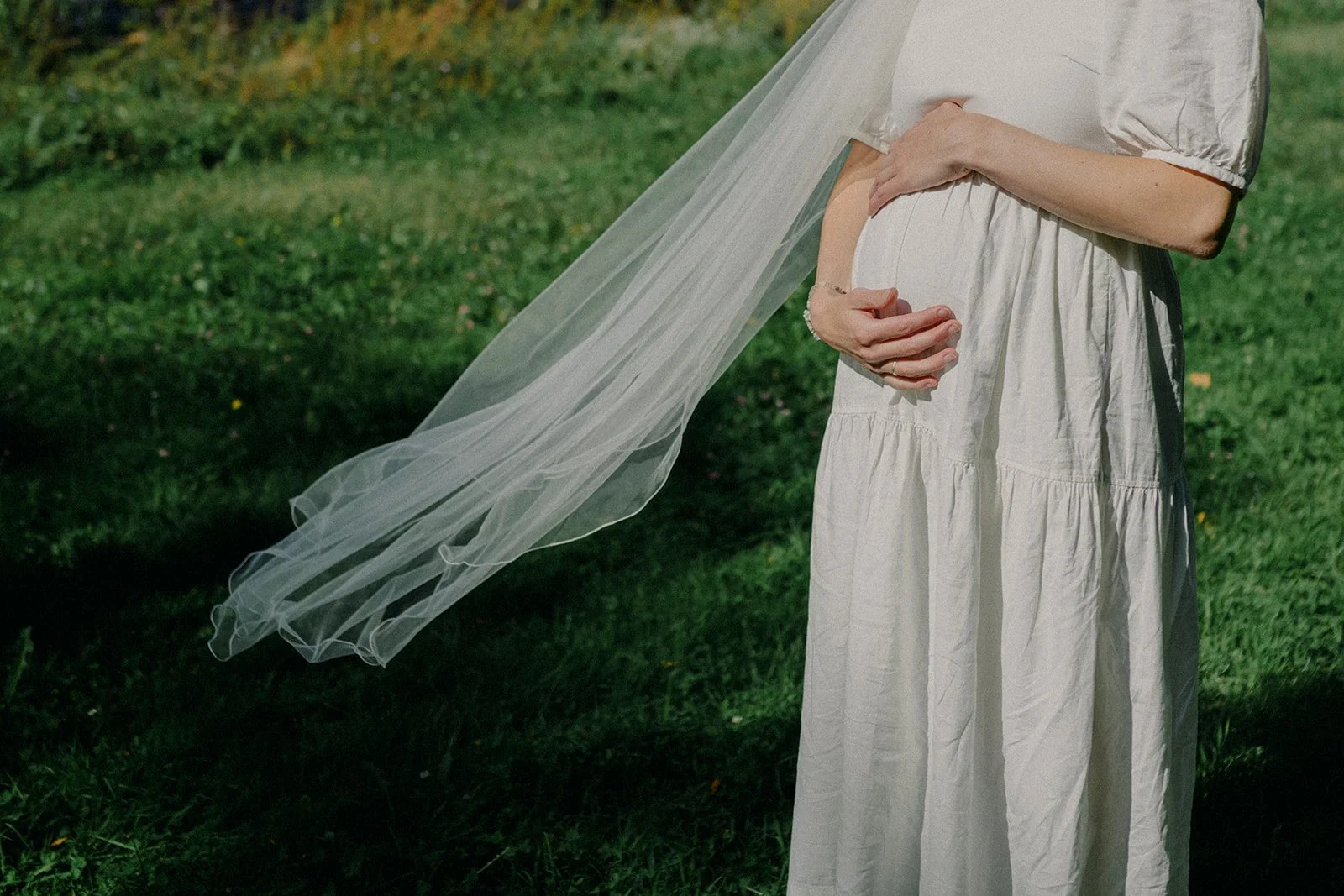 Close-up of a pregnant woman holding her belly outdoors, wearing a white dress and a sheer veil.