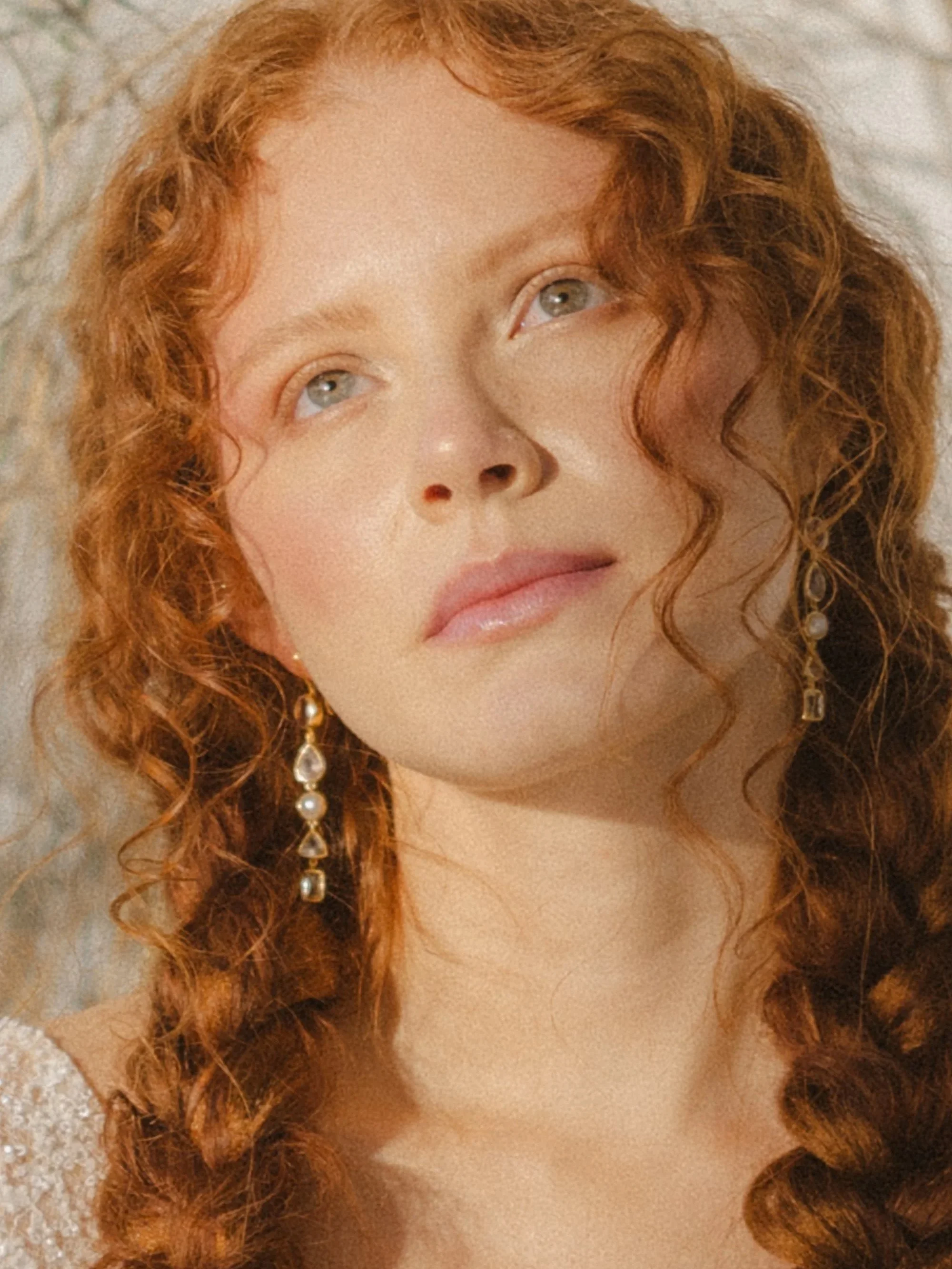Close-up of a woman with curly red hair and blue eyes, wearing pearl and gold earrings, gazing upward with a soft expression.