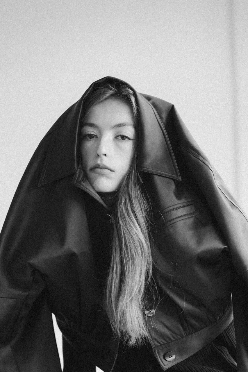 Black and white portrait of a woman with long hair, wearing a leather jacket with the collar up, posing indoors.