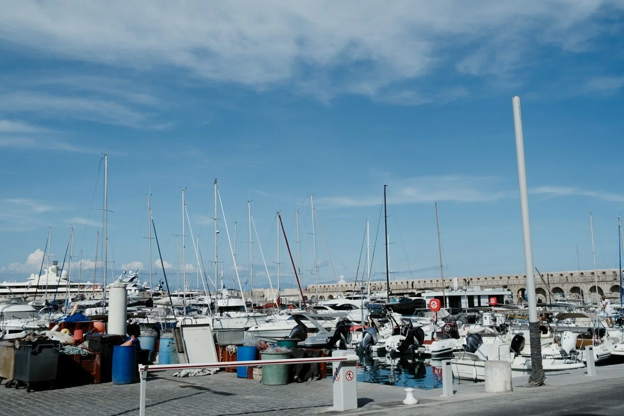 A marina with multiple sailboats and yachts docked, blue sky with clouds, stone pier in the background, some equipment and barrels on the dock.