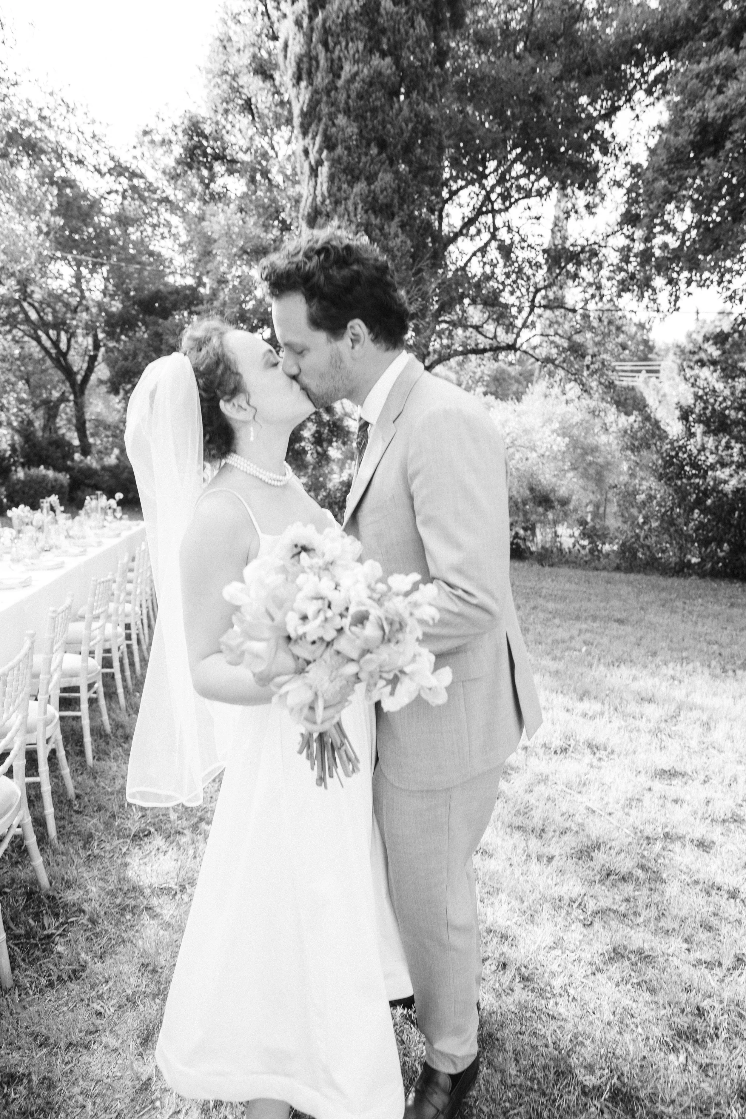 Black and white photo of a bride and groom kissing outdoors, with wedding guest tables in the background.