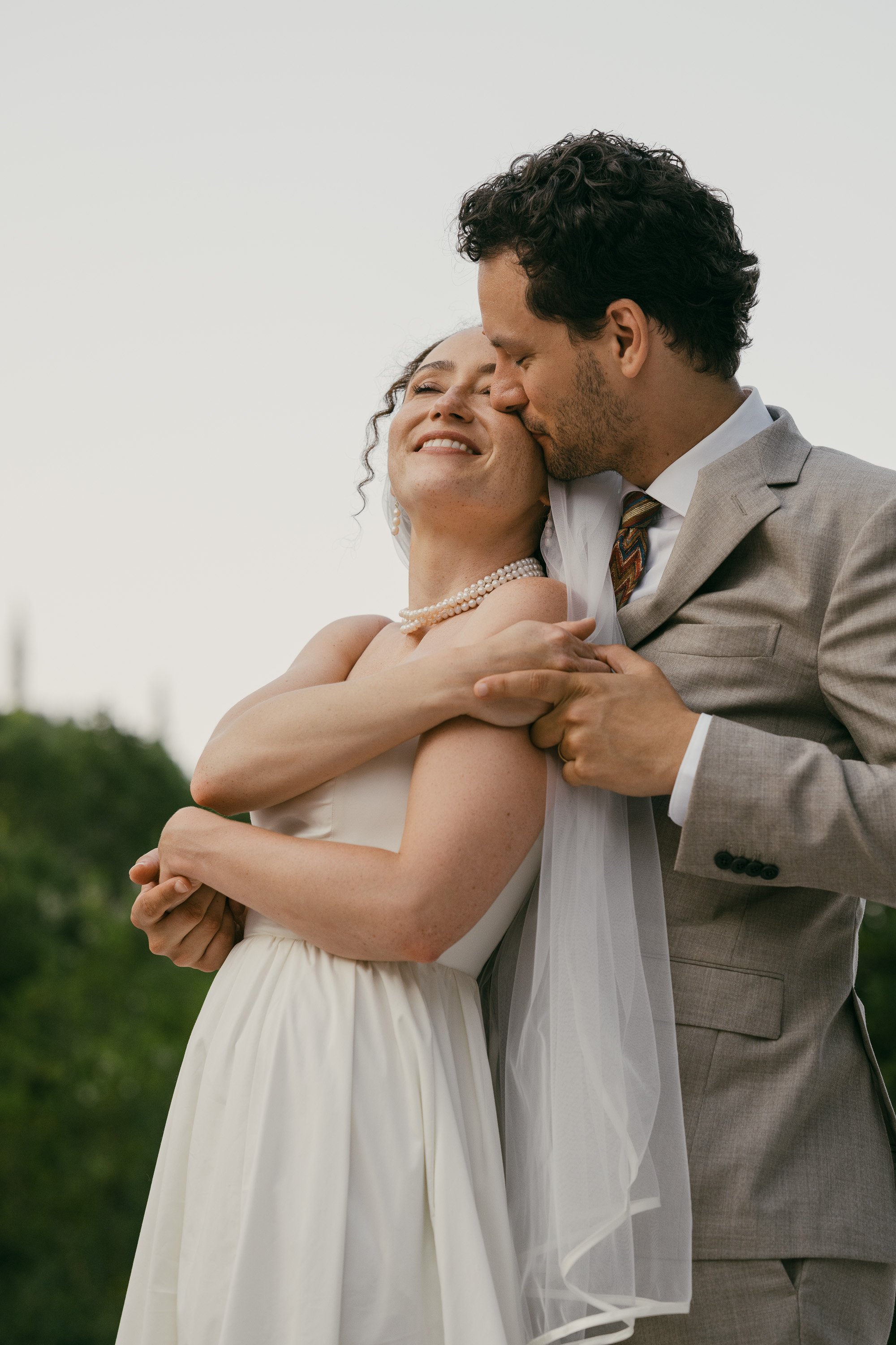 A happy woman in a wedding dress and pearl necklace embracing a man in a grey suit and tie, who is kissing her on the cheek outdoors during the daytime.