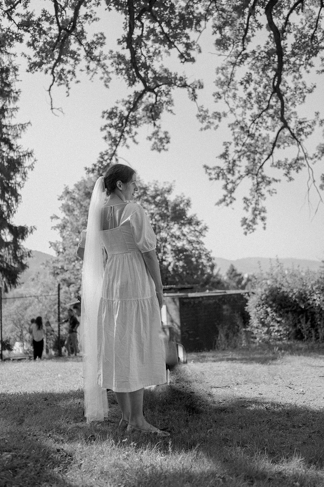 A woman in a wedding dress with a veil standing outdoors under tree branches, looking to her side, with a small group of people and a fenced area in the background.