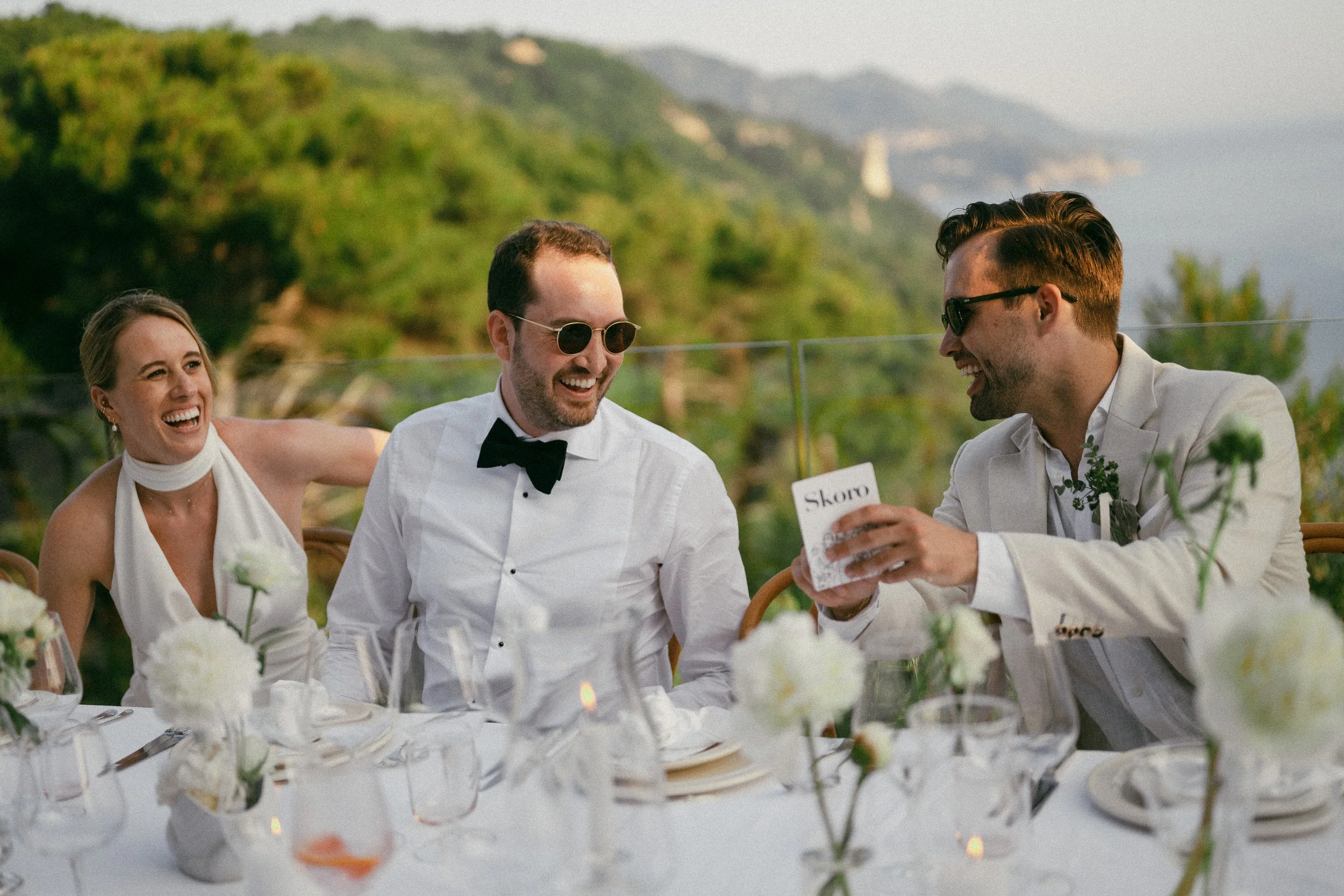 Four people in formal attire sitting at a decorated outdoor table, laughing and smiling, with a scenic green landscape and cliffs in the background during daytime.
