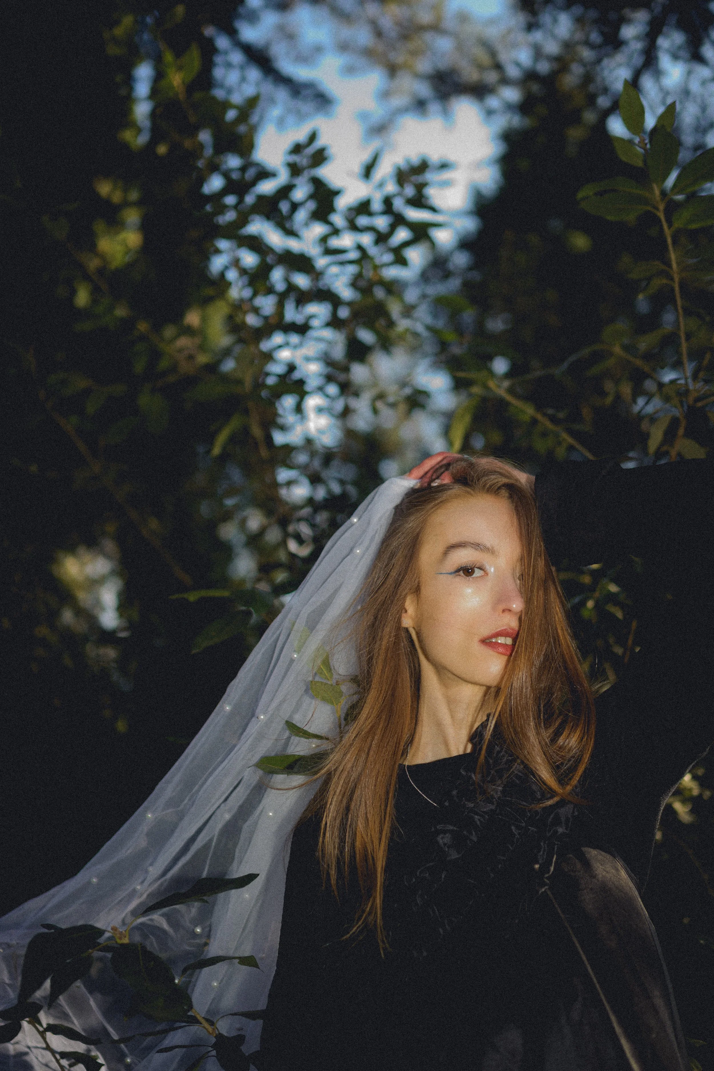 A woman with red hair wearing a white veil and black top, sitting among green leaves and branches in a natural outdoor setting.