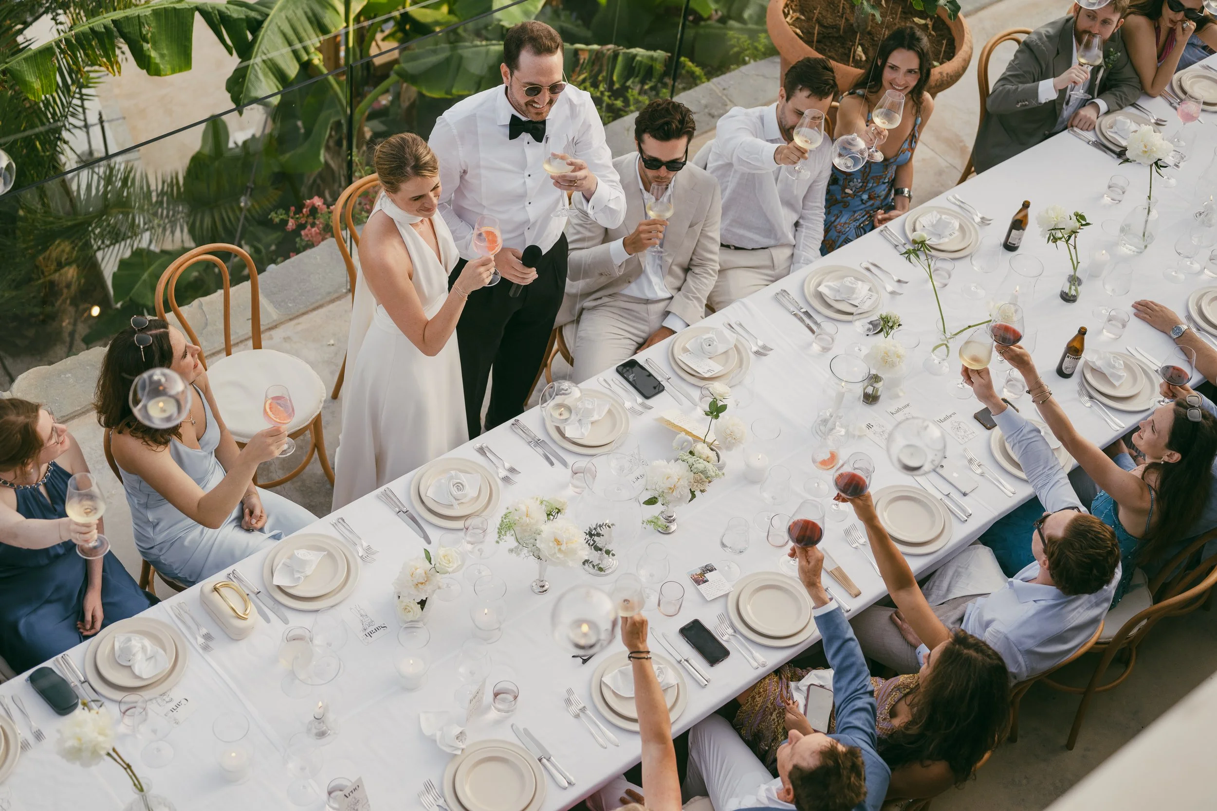 A group of people sitting and standing around a long table outdoors, raising glasses in a toast. The table is decorated with white flowers and white plate settings. The setting appears to be a celebration or wedding reception with a greenery backdrop