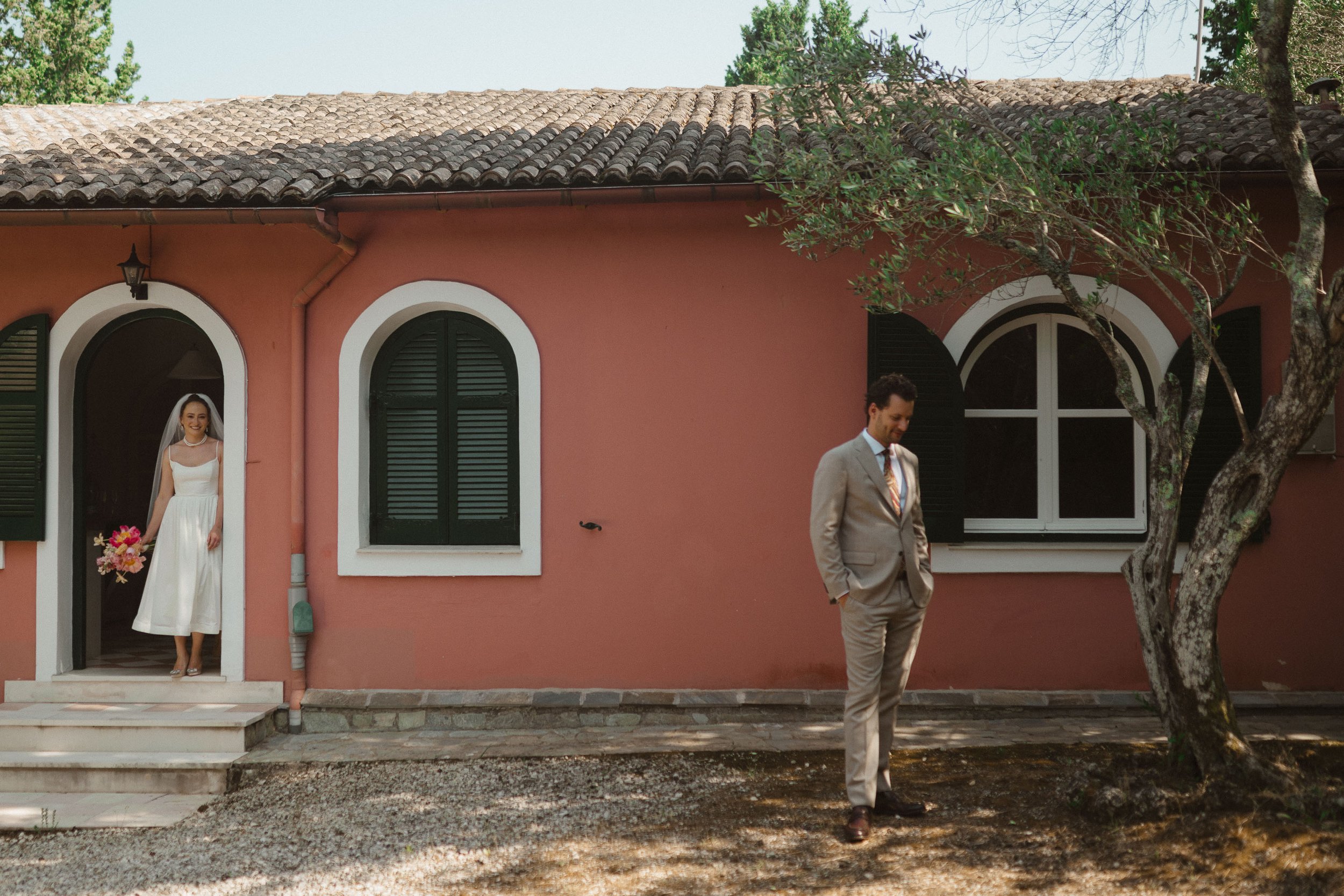 A bride in a white dress holding a bouquet stands in a doorway, smiling, while a groom in a gray suit stands outside by a tree, looking down with hands in pockets, in front of a pink house with green shutters.