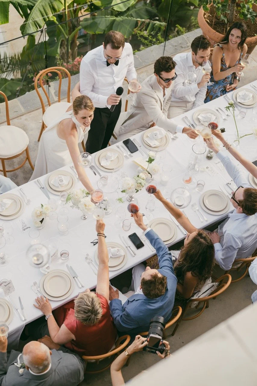A wedding reception with a bride in a white dress and groom in a beige suit seated at a long outdoor dining table with friends and family. People are raising glasses of wine in a toast, and one person is standing with a microphone. The table is decor