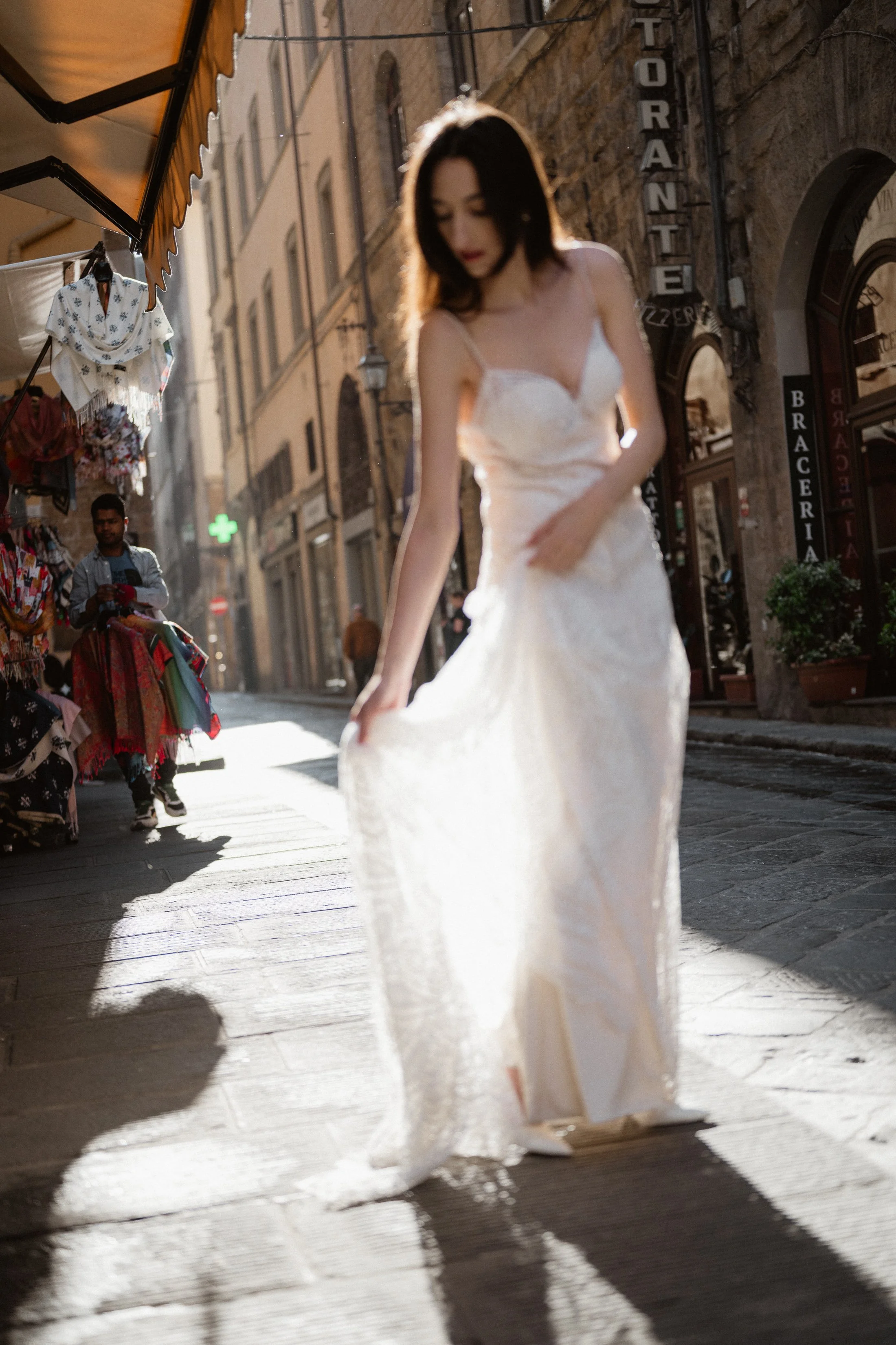 A woman in a white dress walking on a sunlit cobblestone street in a European city, with storefronts and a street vendor nearby.