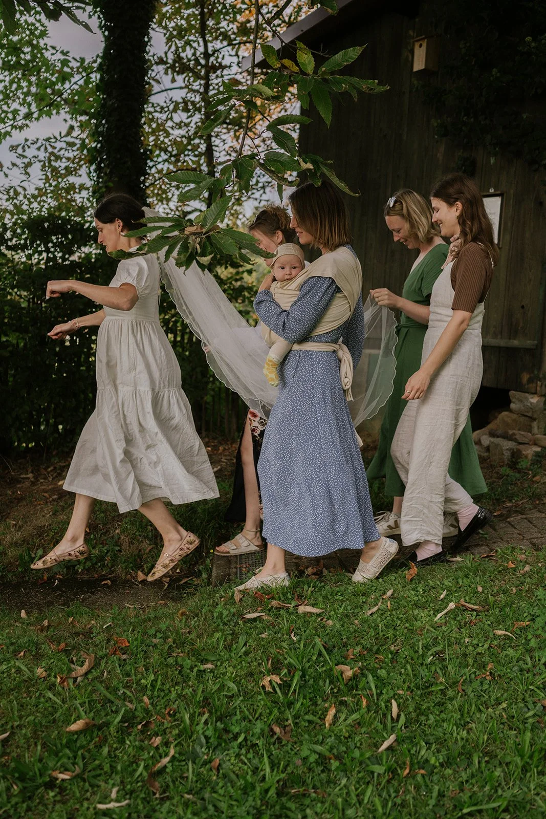 Group of women walking in a line outdoors, one holding a small child, under a tree with green leaves, on a grassy area.
