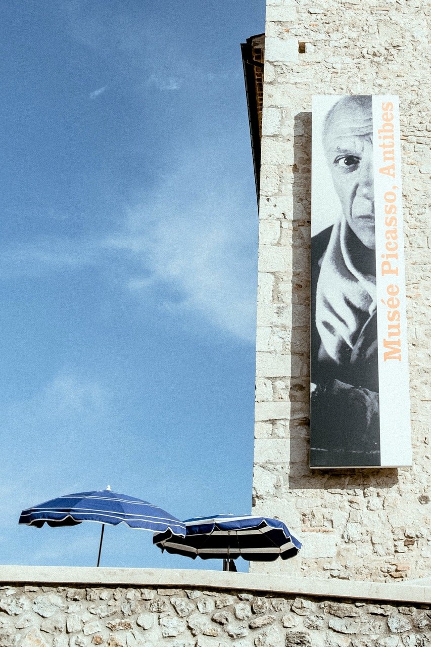A large banner on an old stone building featuring a black and white picture of Picasso's face and the text 'Musée Picasso, Antibes'. Two blue and white striped umbrellas are on a stone balcony below the banner against a blue sky.
