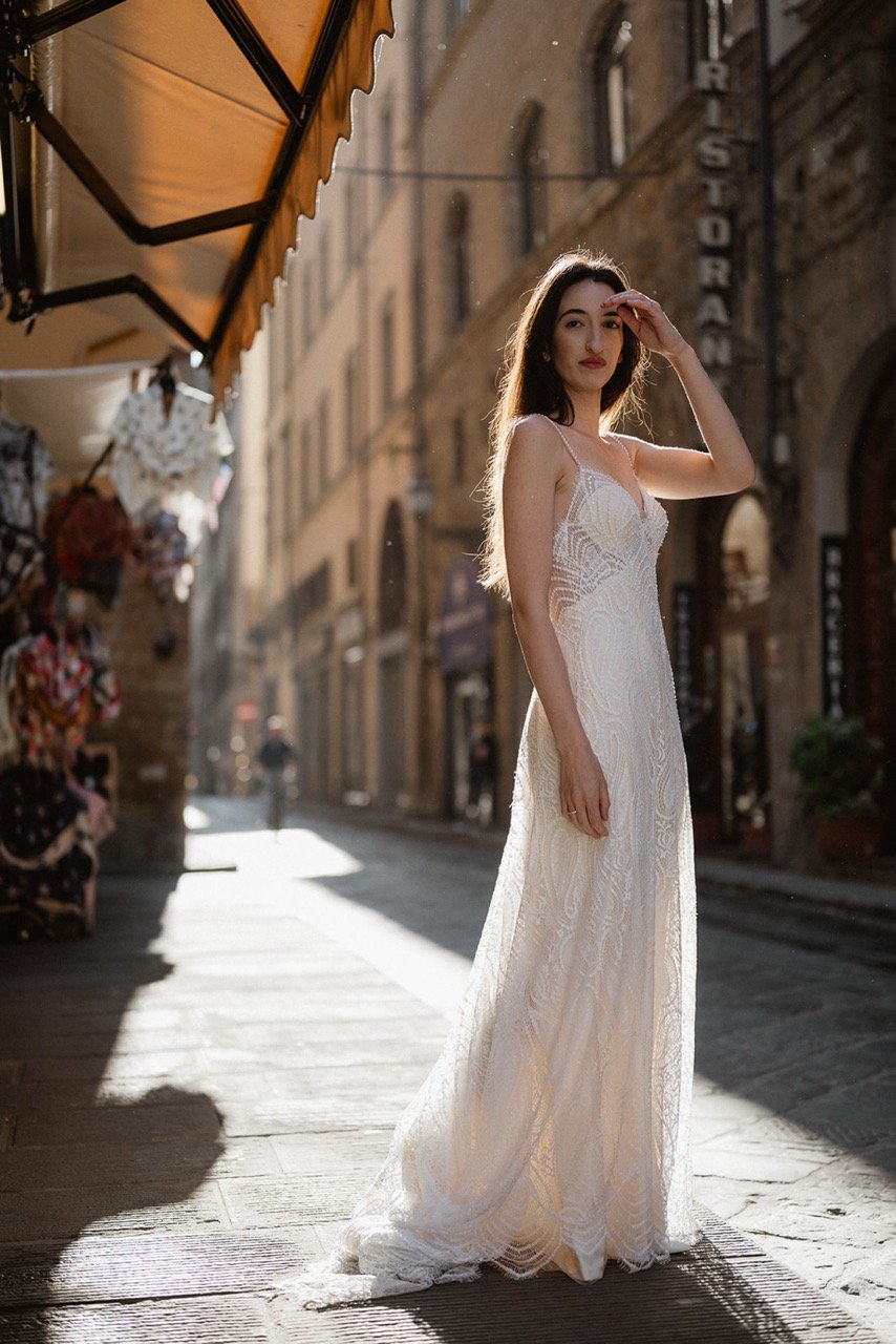 A young woman in a white lace dress standing on a sunlit city street, shielding her eyes with her hand.