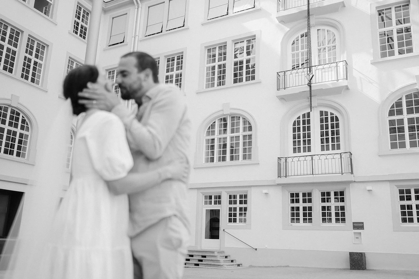 A black and white photo of a couple in a romantic embrace, kissing, in front of a modern white building with large windows and balconies.