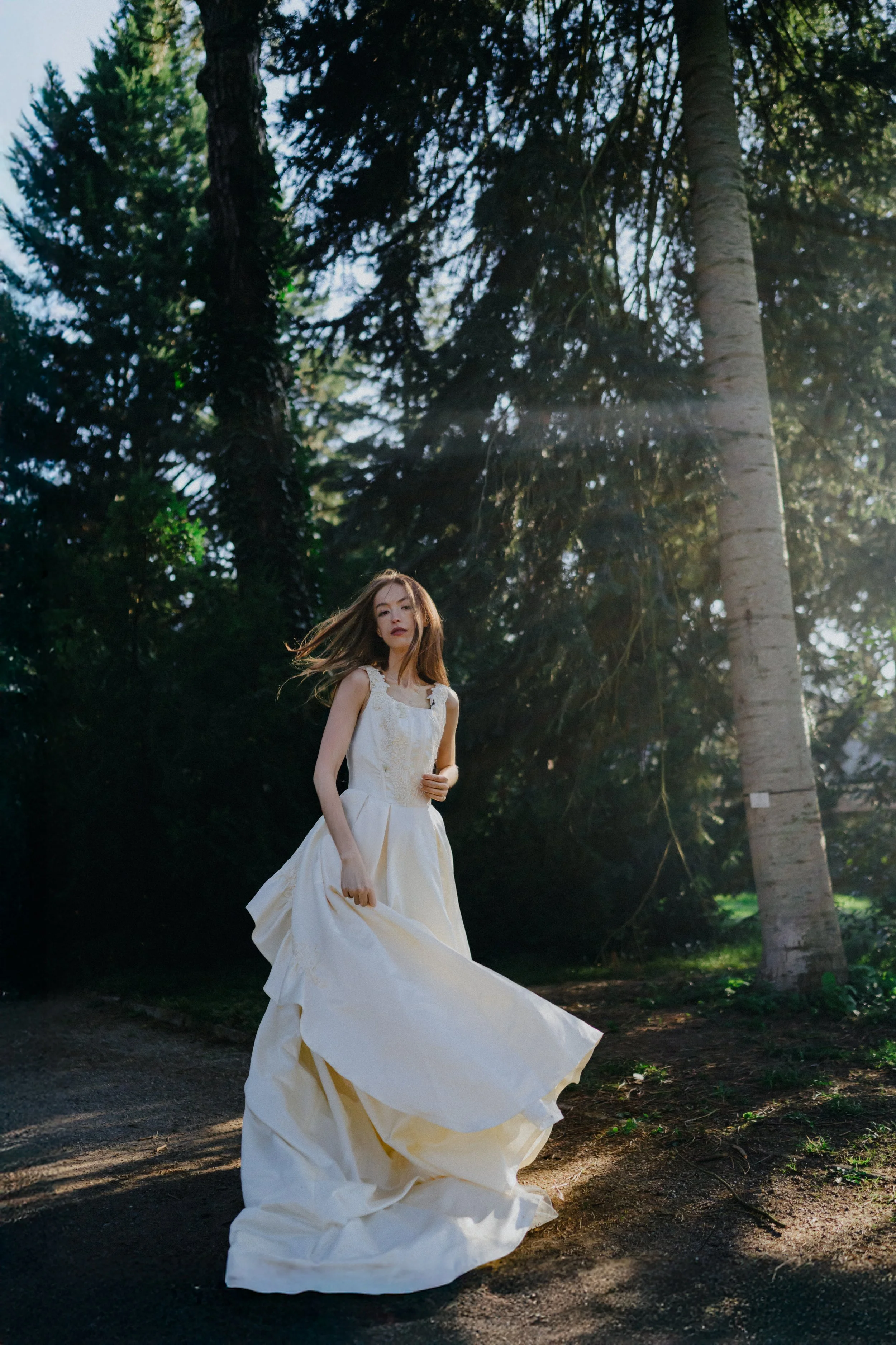A woman in a flowing white dress standing on a dirt path in a forest, with tall trees and sunlight filtering through the foliage.