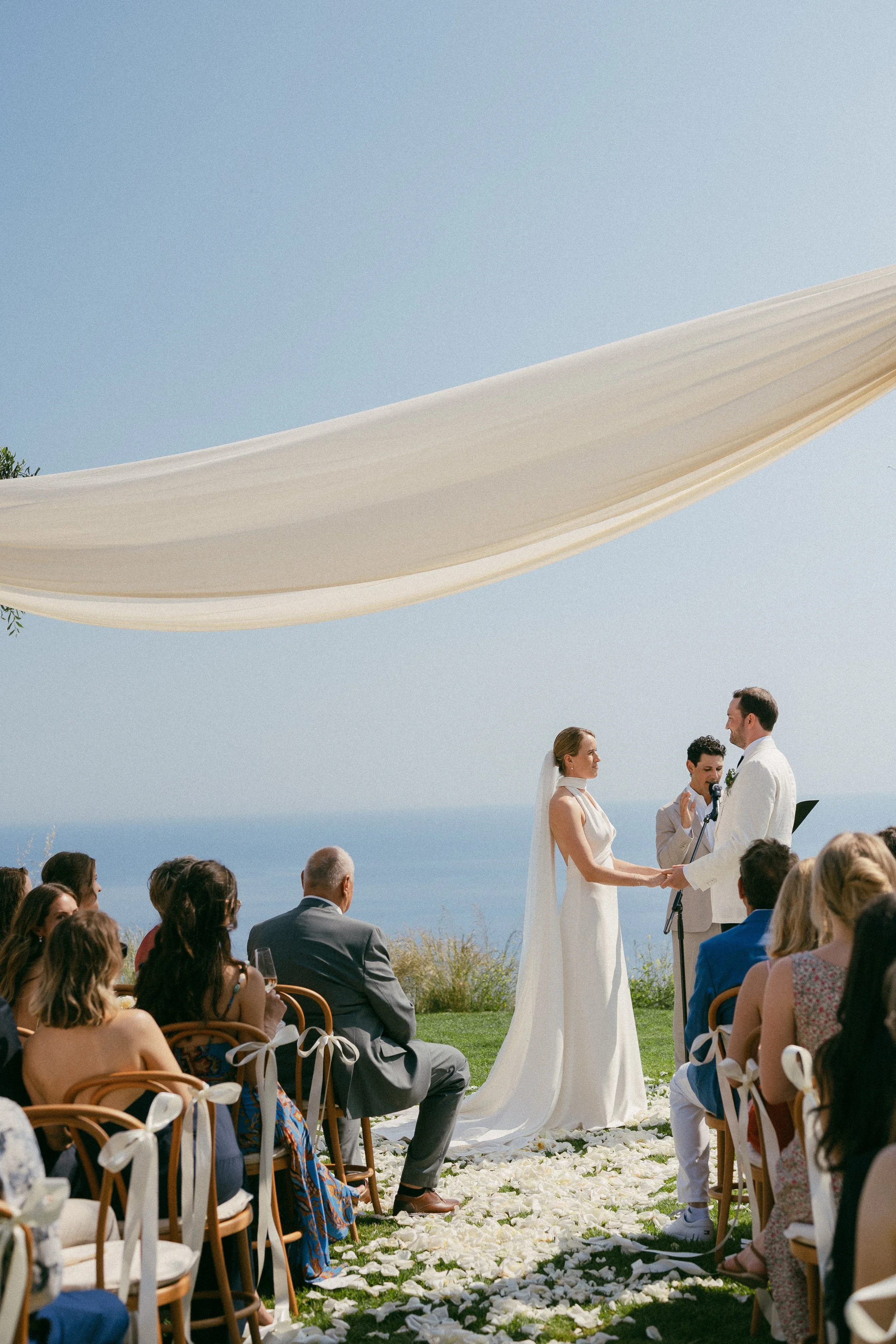 A wedding ceremony taking place outdoors by the coast, with a bride and groom holding hands and exchanging vows under a white canopy, surrounded by seated guests.