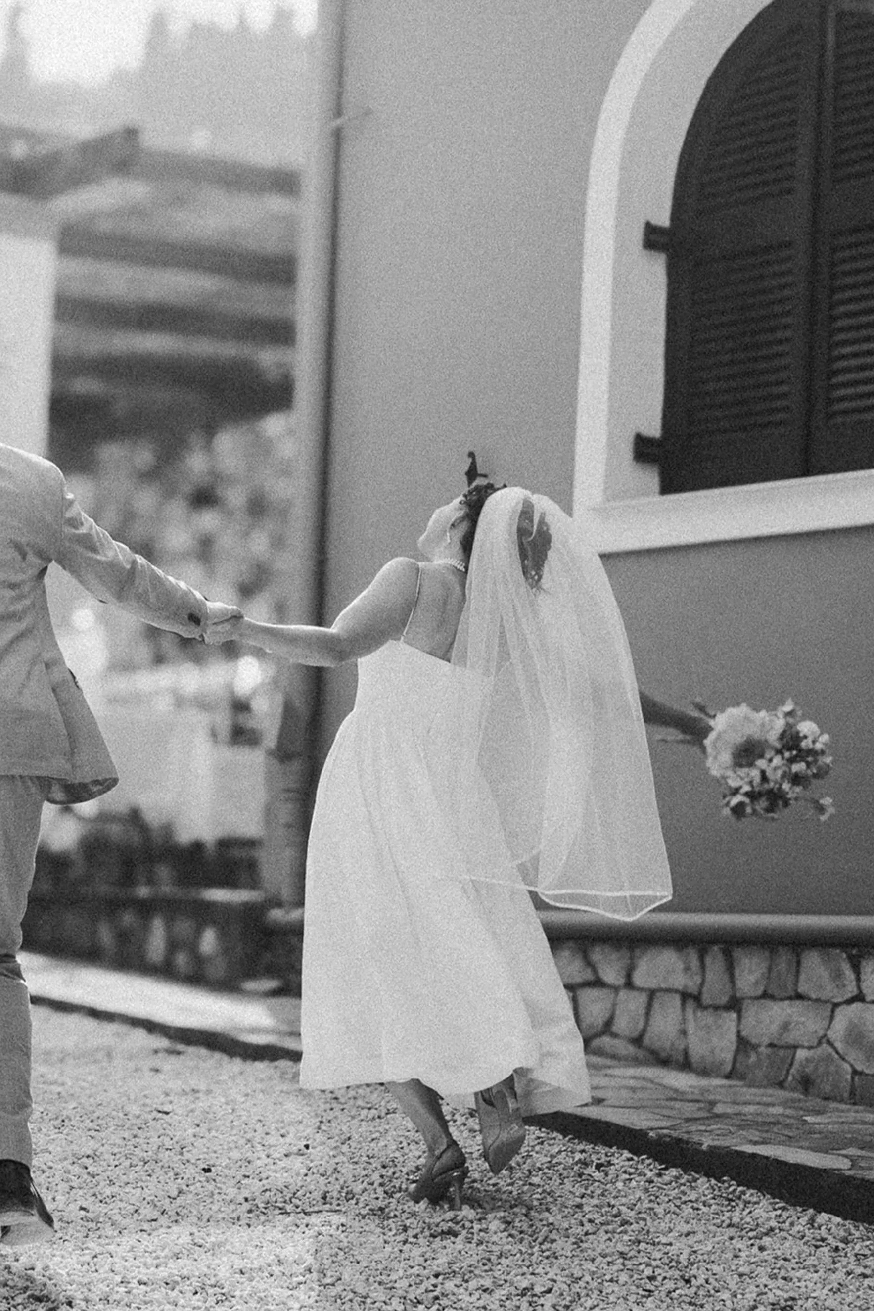 A bride and groom holding hands and dancing outdoors, with the bride wearing a wedding dress and veil, and the groom in a suit, in front of a building with arched windows and shutters.