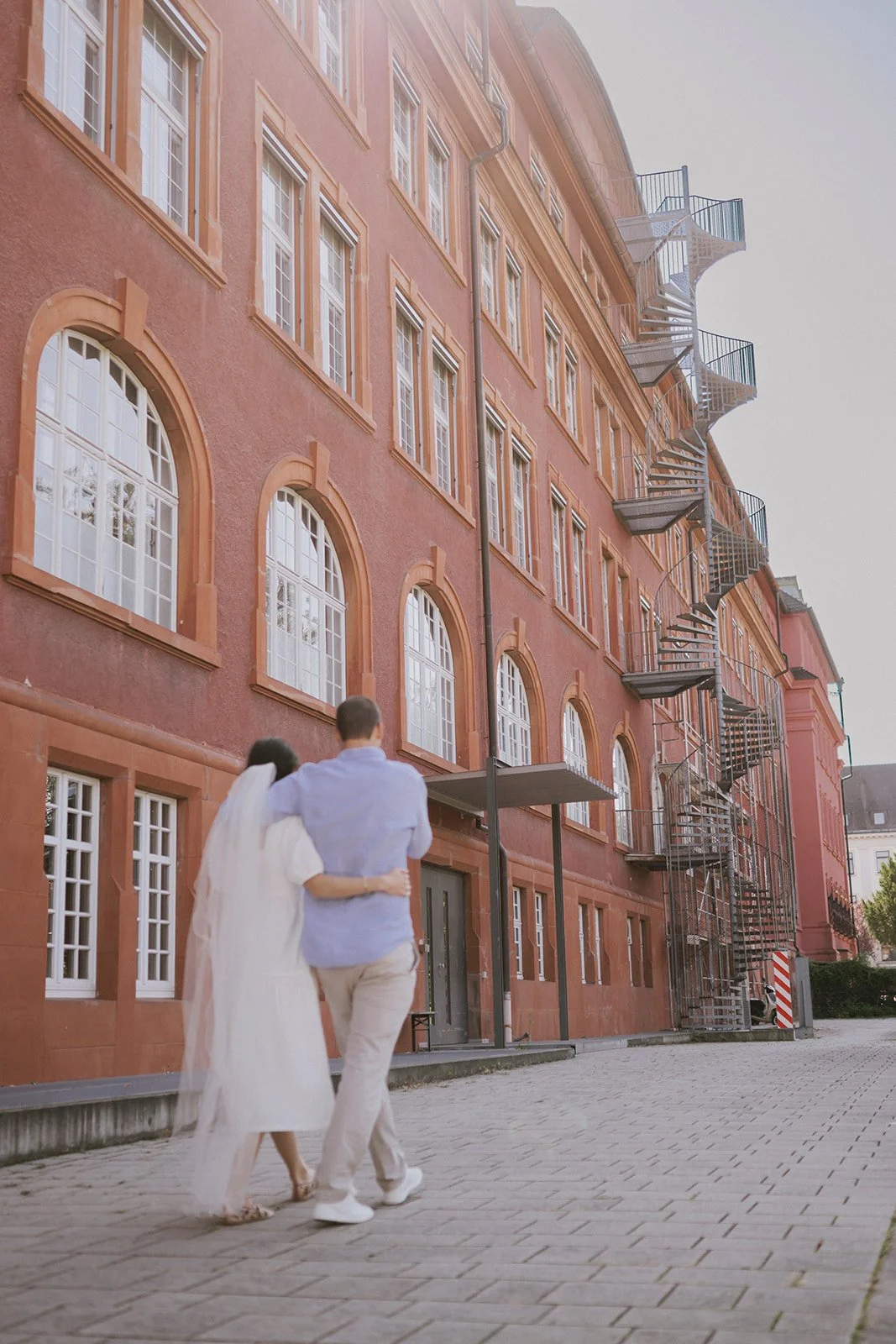 A couple walking arm-in-arm in front of a pink multi-story building with large arched windows and an external fire escape stairs.