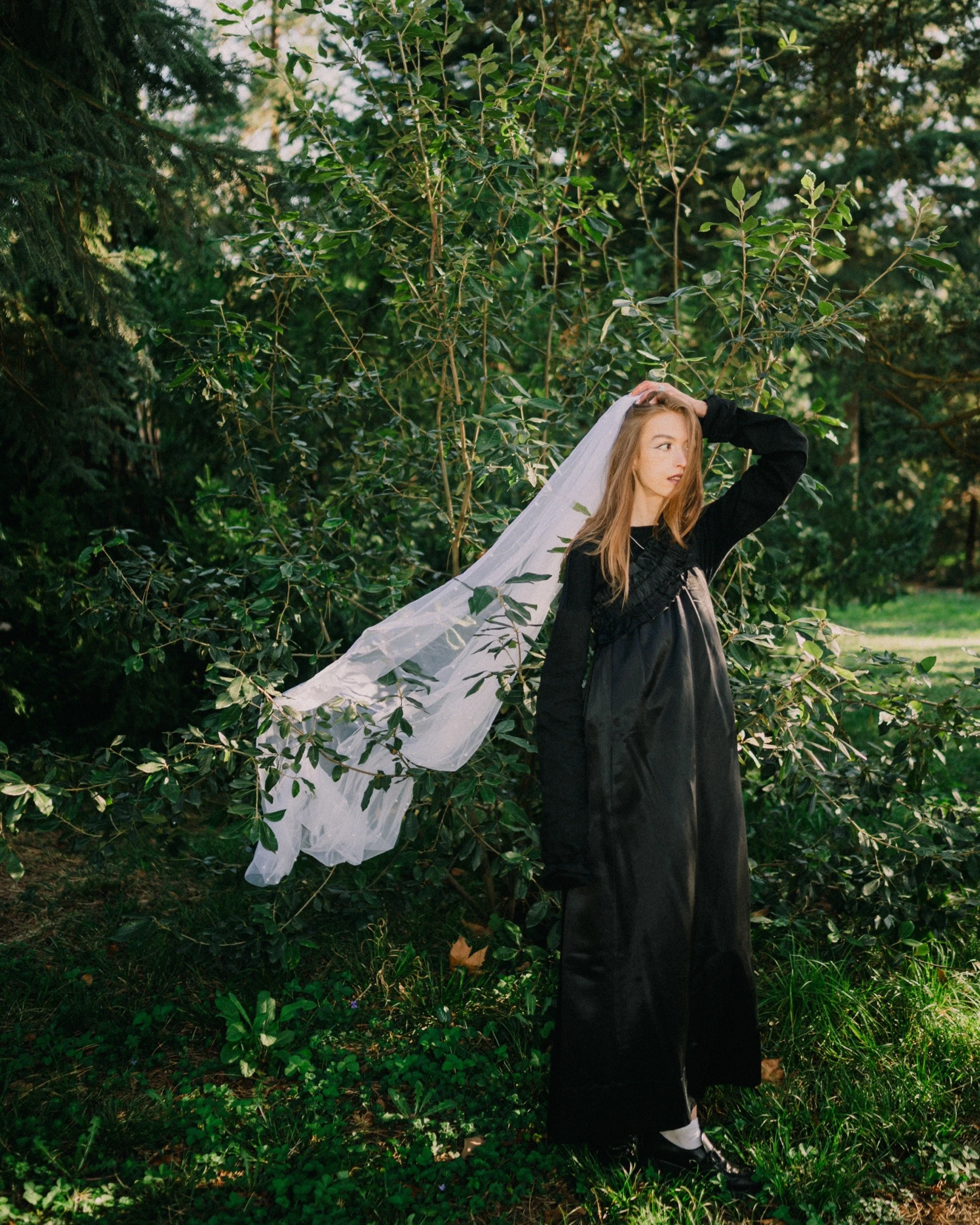 Young woman in black dress with a sheer veil standing outdoors among greenery and trees