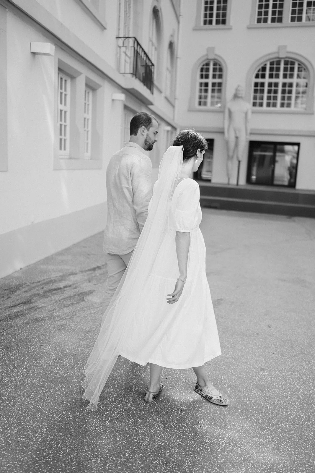 A black and white photo of a bride and groom walking outside, with her wedding dress and veil, in front of a building with tall windows and a sculpture of a human figure.