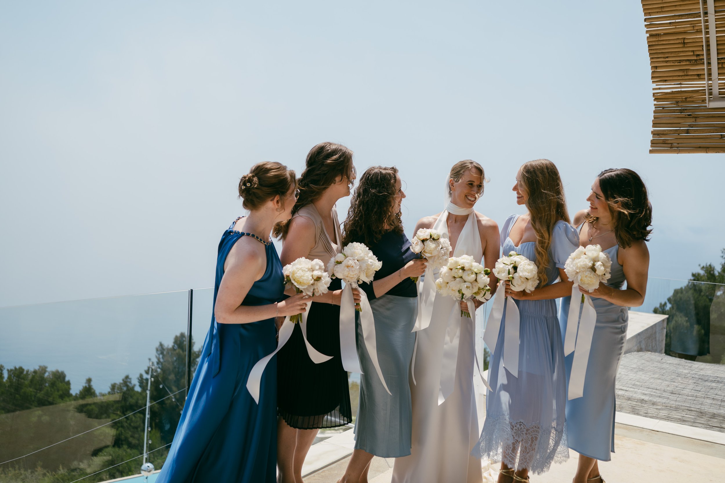 Group of bridesmaids and bride at a wedding, holding white bouquets and smiling on an outdoor terrace with a glass railing and scenic landscape in the background.