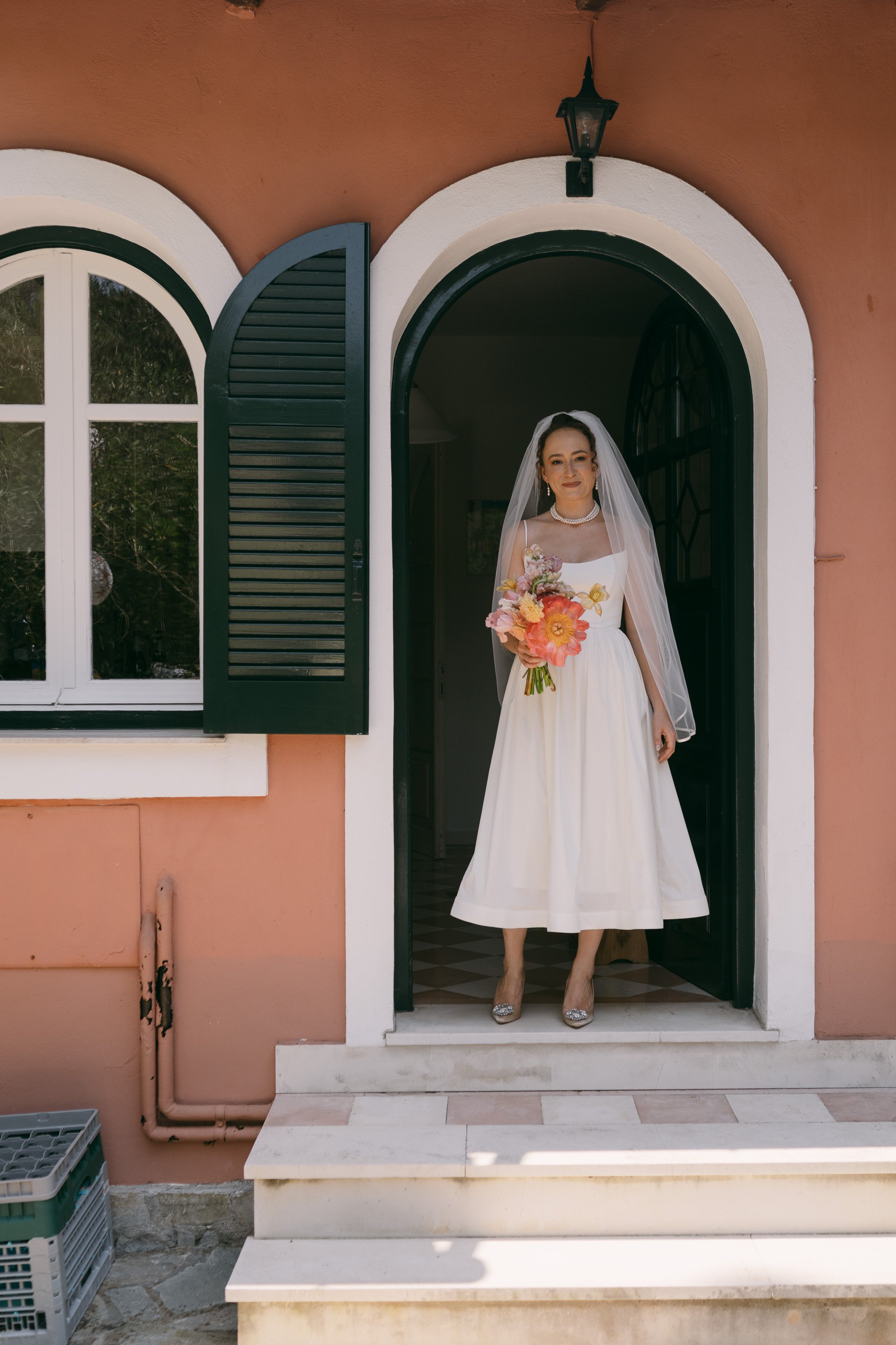 A woman in a white wedding dress holding a bouquet of pink, orange, and purple flowers, standing at the doorway of a house with pink walls, a black door, and an arched window.