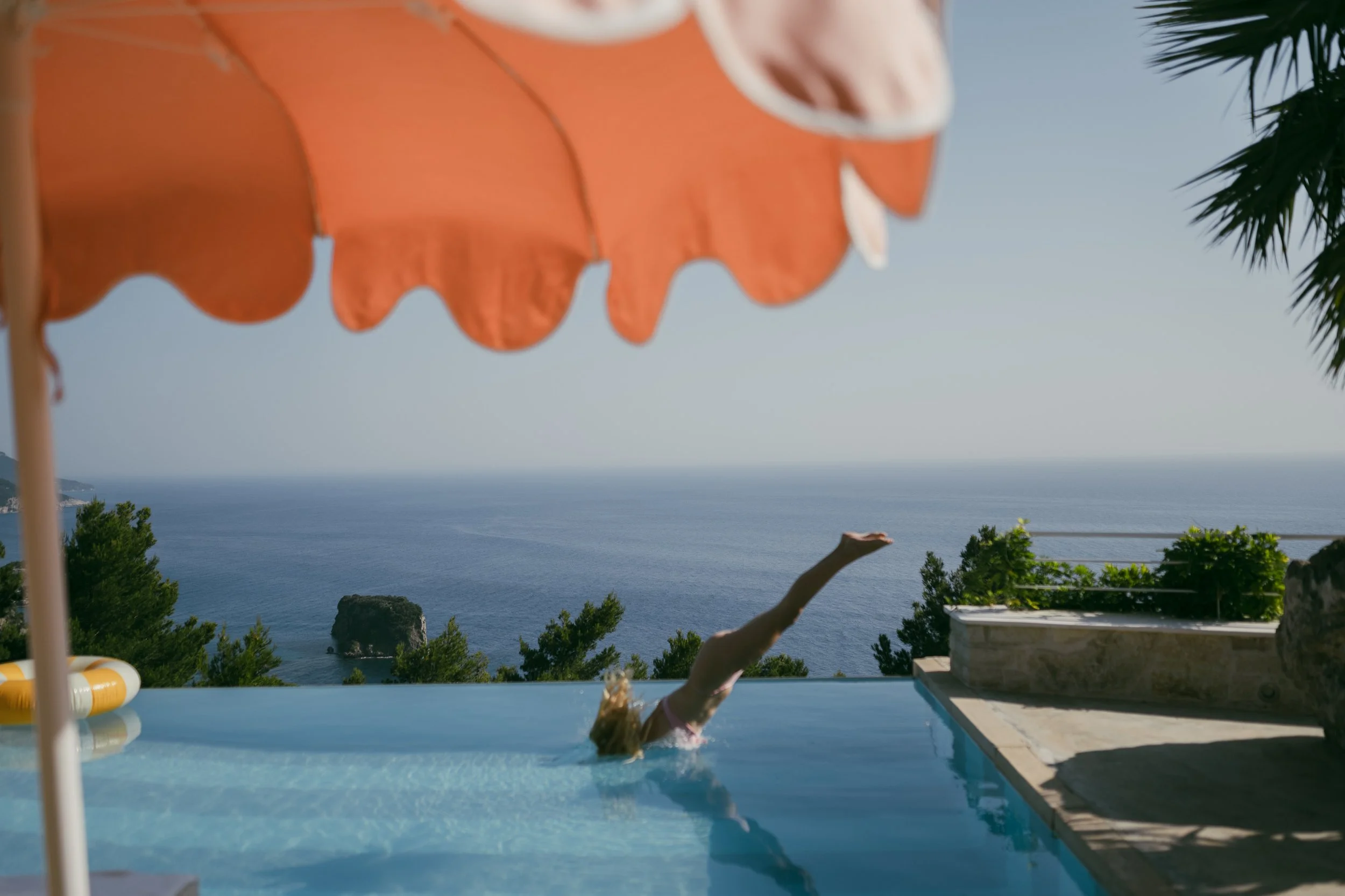 Person diving into an infinity pool overlooking the ocean, with an orange patio umbrella and greenery.