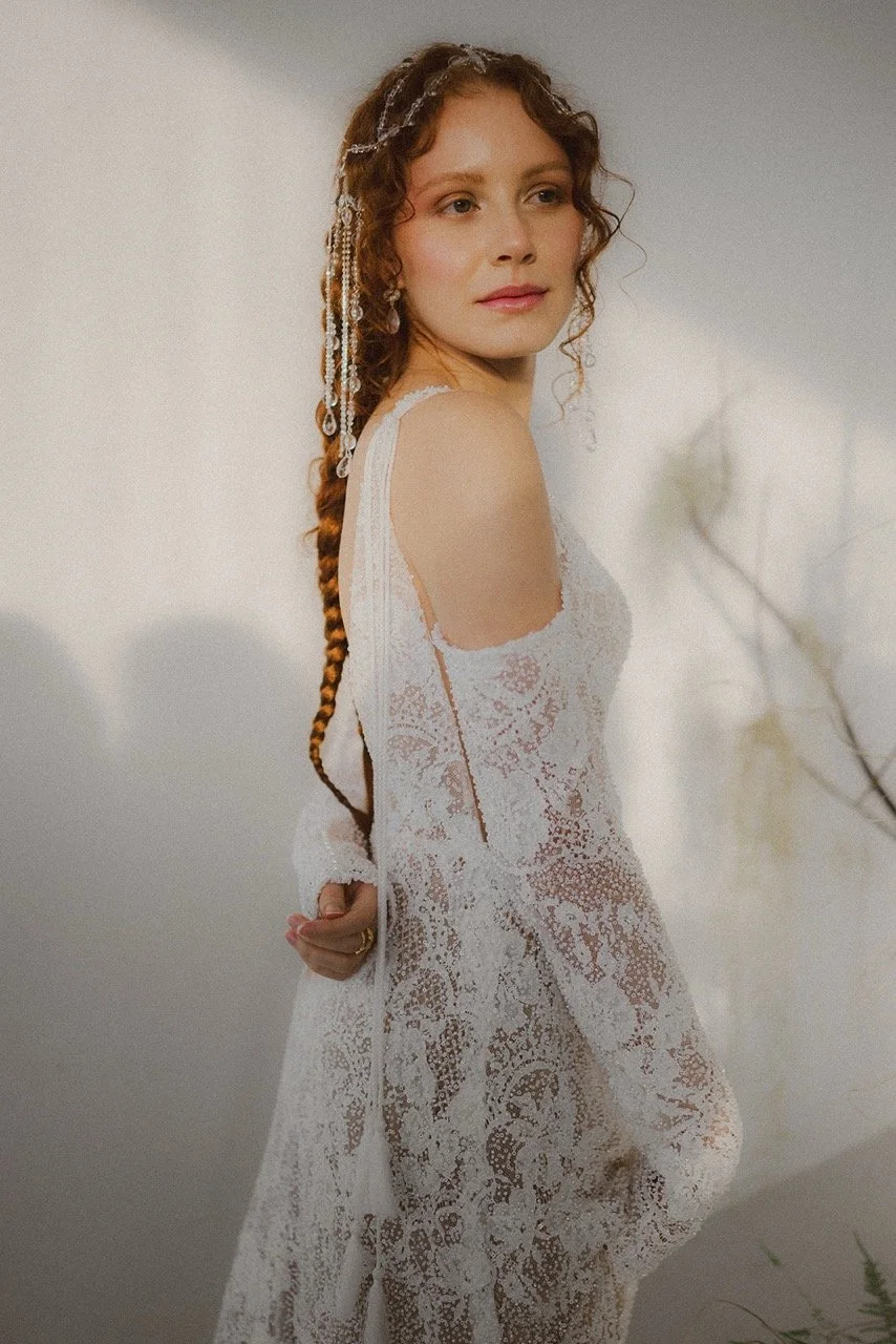 Woman in a white lace wedding dress with cold shoulder sleeves, with long braided hair adorned with pearl and jewelry accessories, looking over her shoulder against a neutral background.