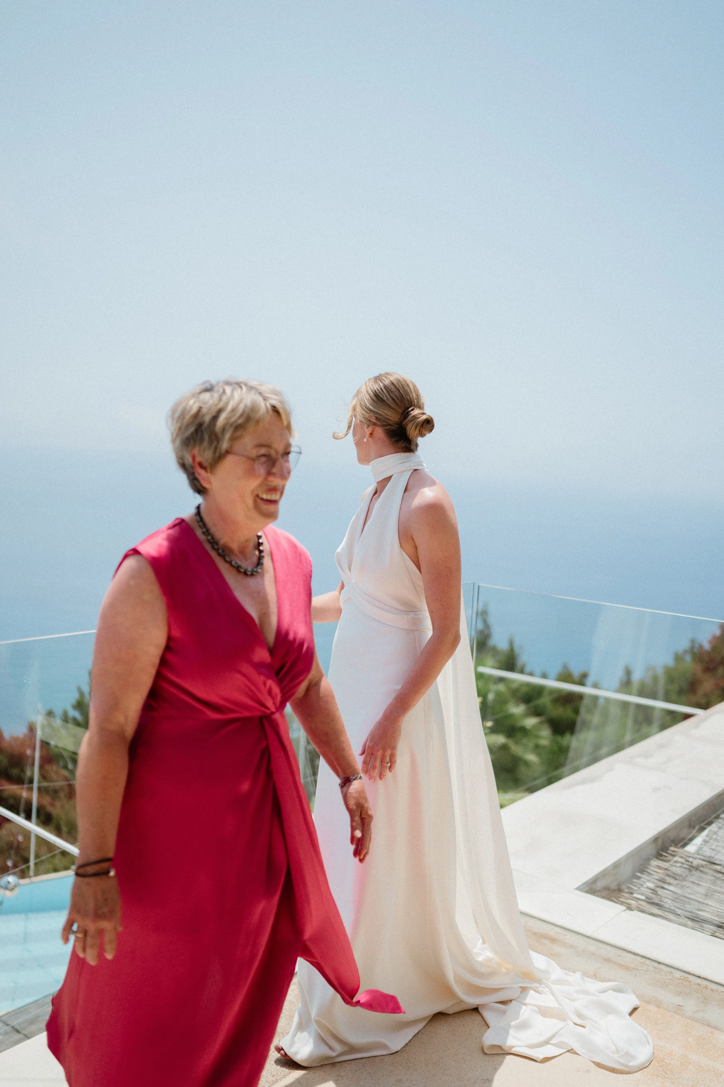 A woman in a white dress and a woman in a red dress standing outdoors on a terrace with a glass railing, overlooking a scenic view of the ocean and sky during the daytime.