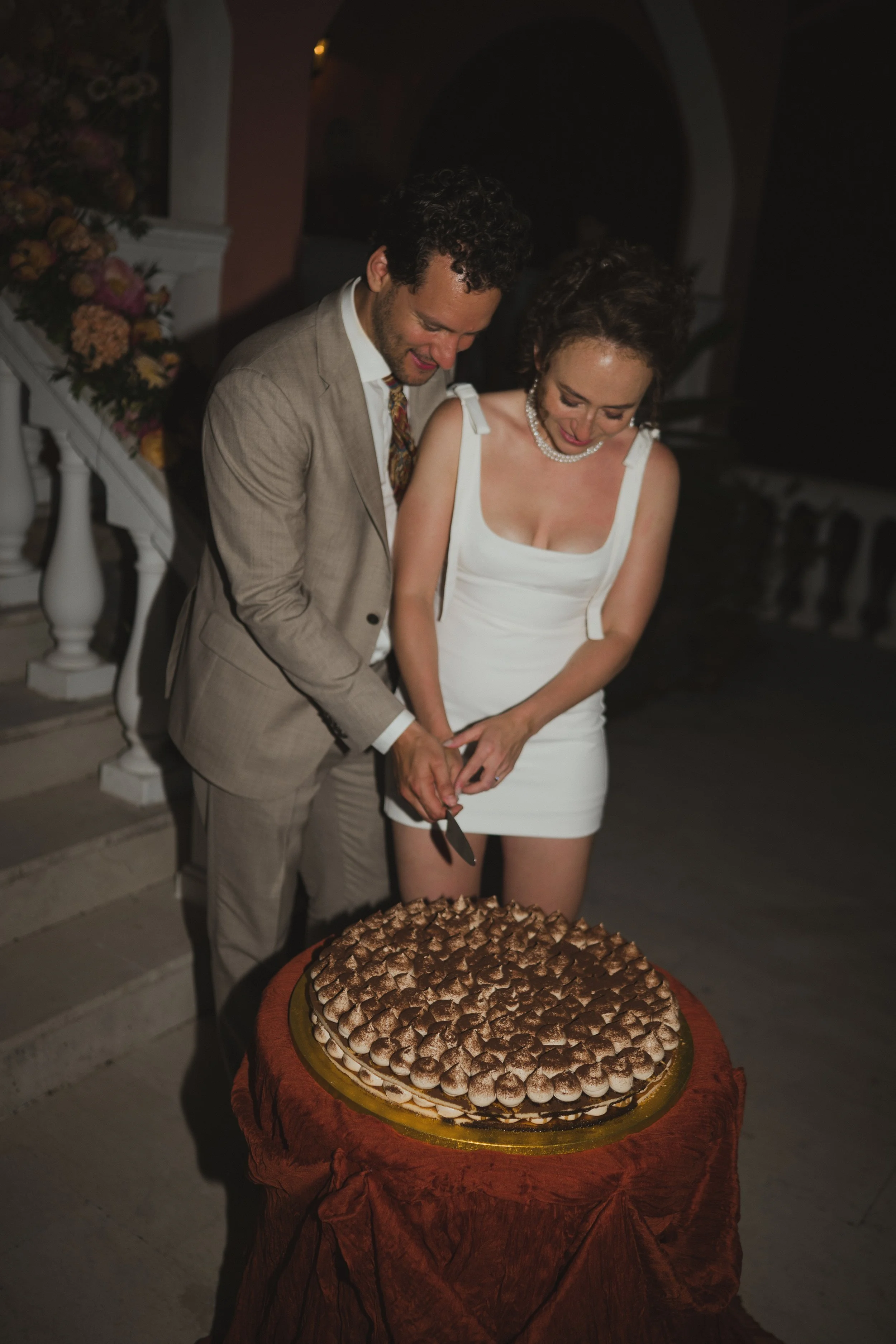 A couple is cutting a large layered cake with chocolate frosting. They are dressed in formal attire, with the woman in a white dress and the man in a beige suit, at a celebration or special event. The cake is on a round table with a brown cloth.