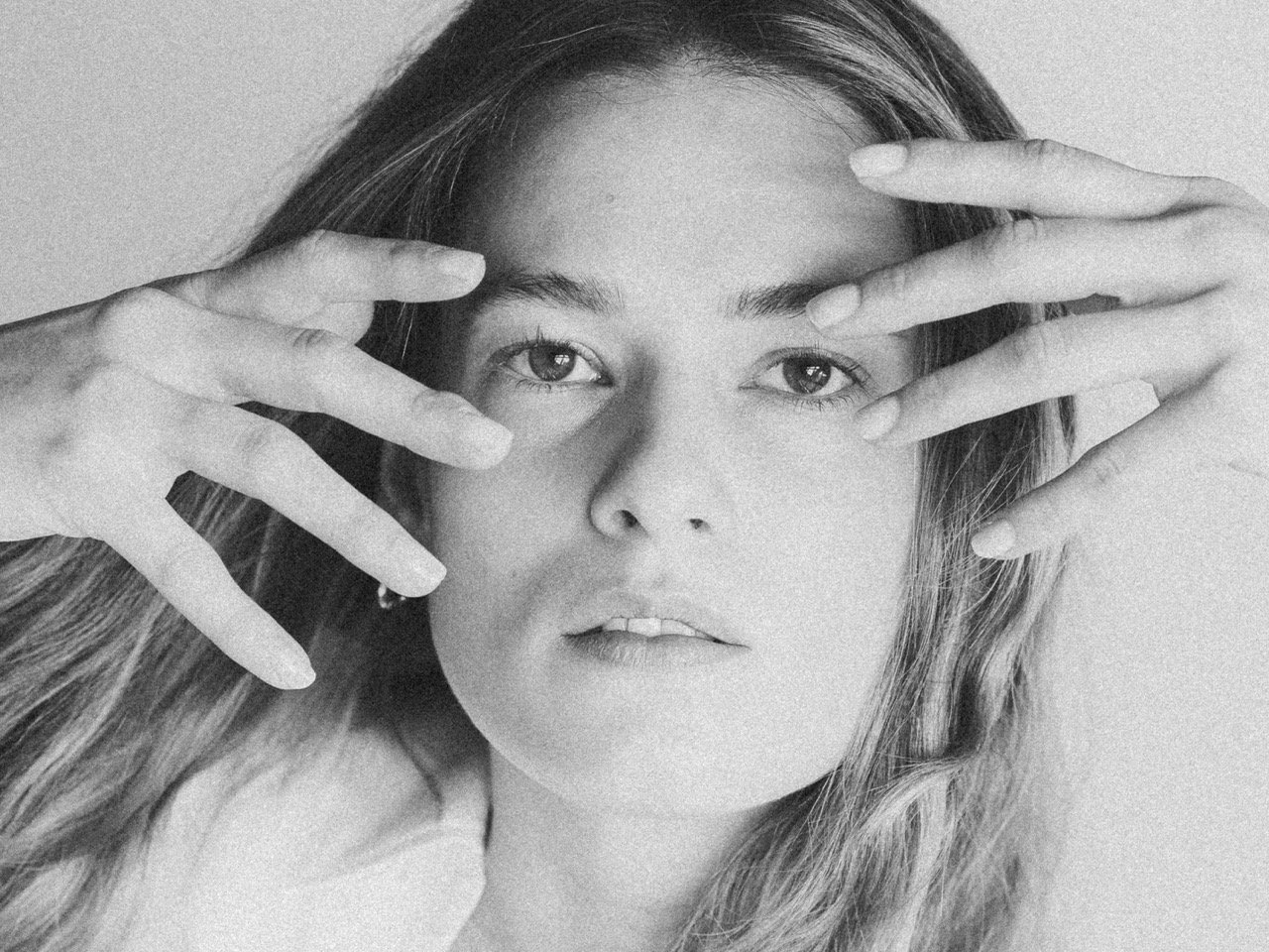 A black and white portrait of a young woman with long wavy hair, placing her hands near her face, with fingers spread around her eyes.