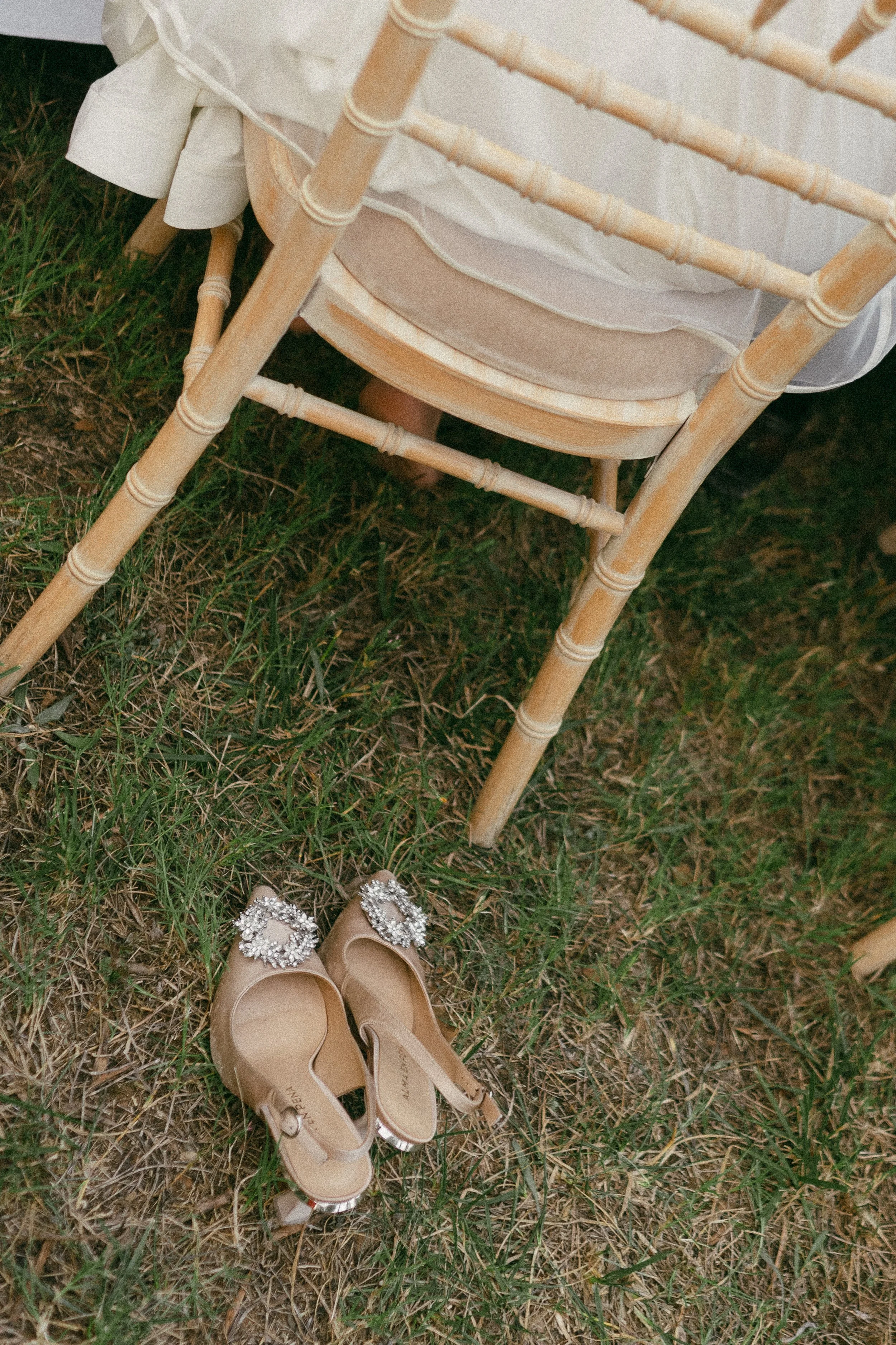 A pair of elegant, beige high-heeled shoes with jeweled embellishments on the toes placed on the grass near a wooden chair with a cushioned seat, part of a table draped with a white tablecloth.