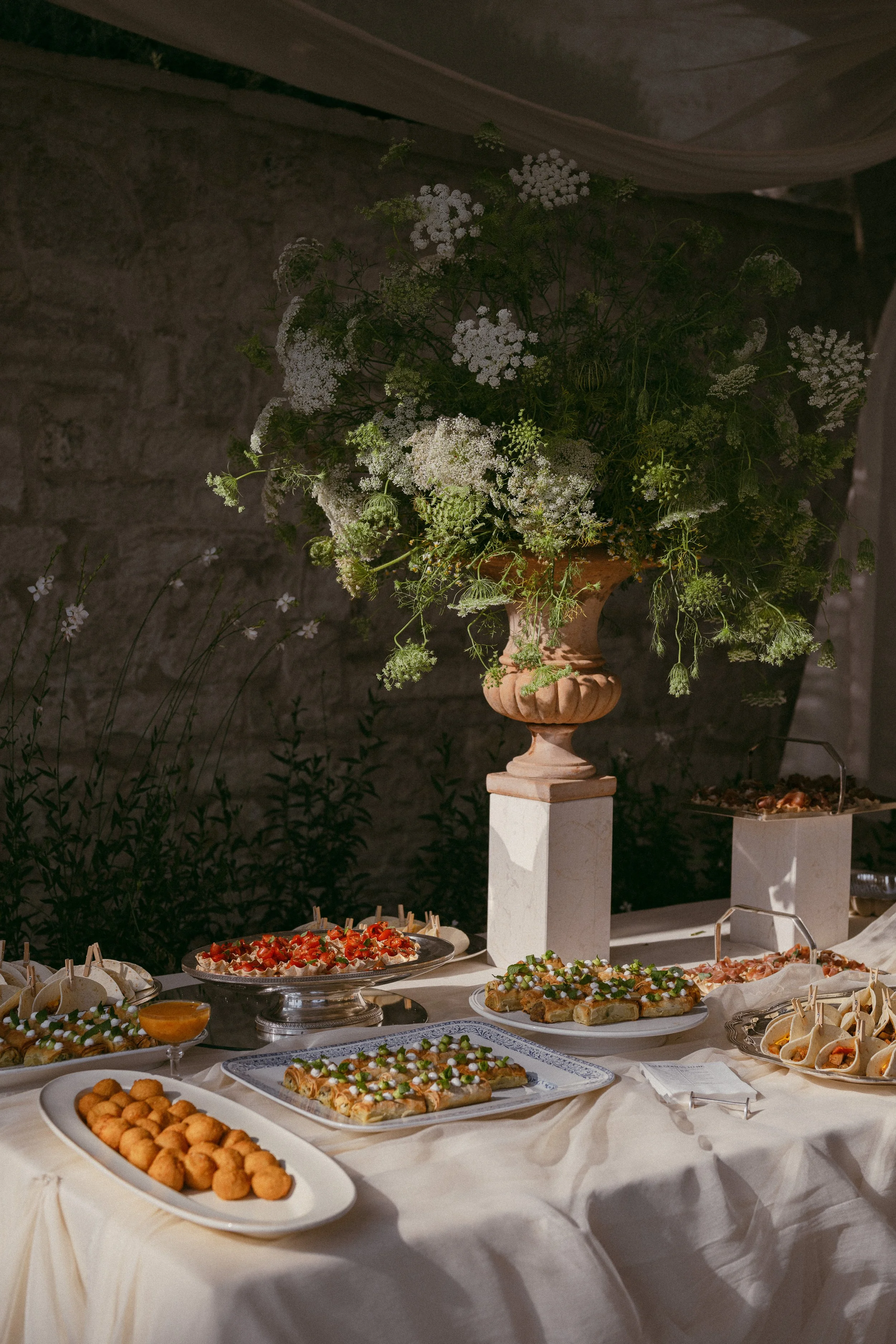 A table with an elaborate dessert spread featuring various pastries, finger foods, and appetizers arranged on silver and white platters, with a large floral arrangement of white flowers in a decorative urn as a centerpiece.