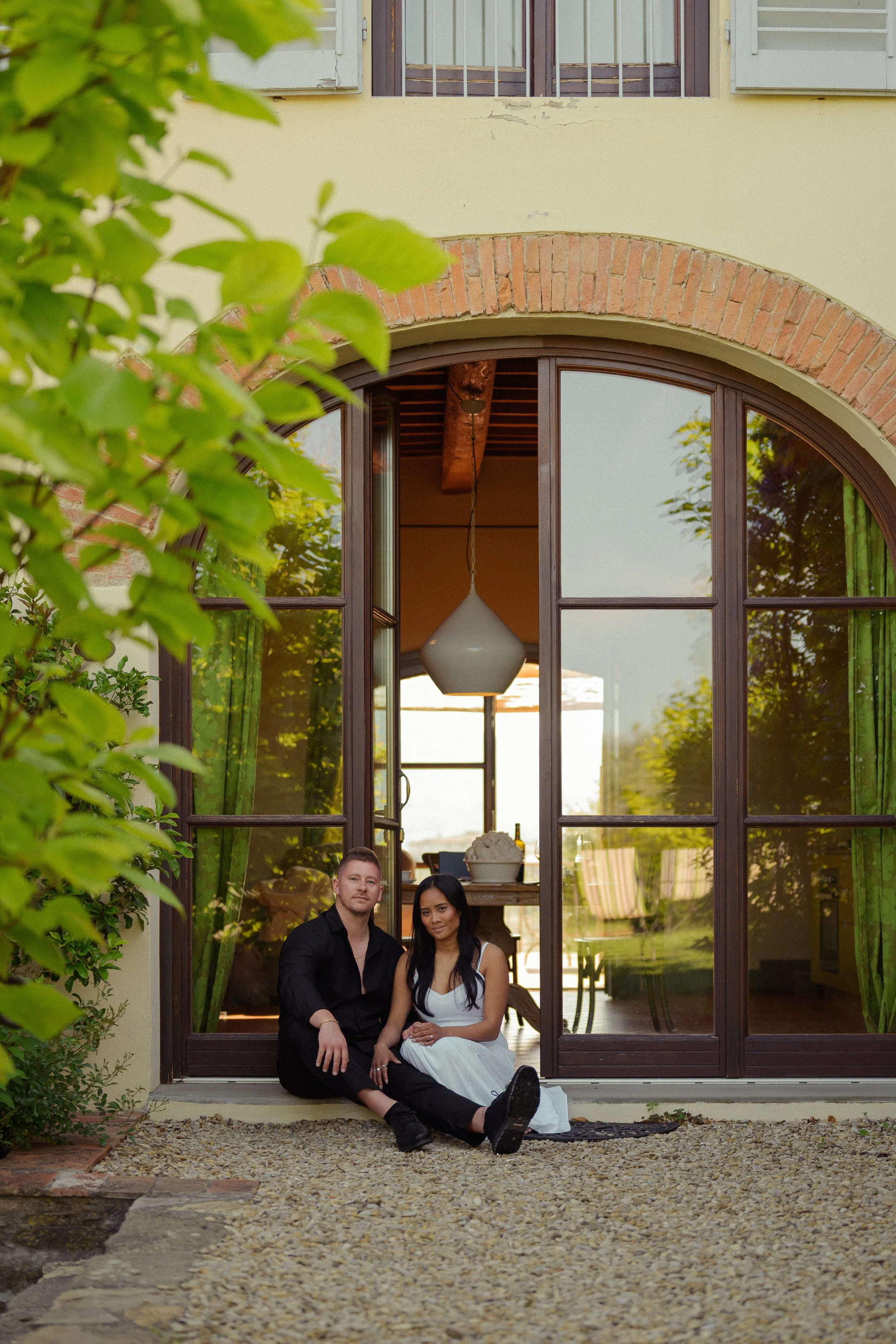 A couple sitting on a gravel patio outside a house with large glass doors, green curtains, and a hanging white light fixture, with trees reflecting in the glass.