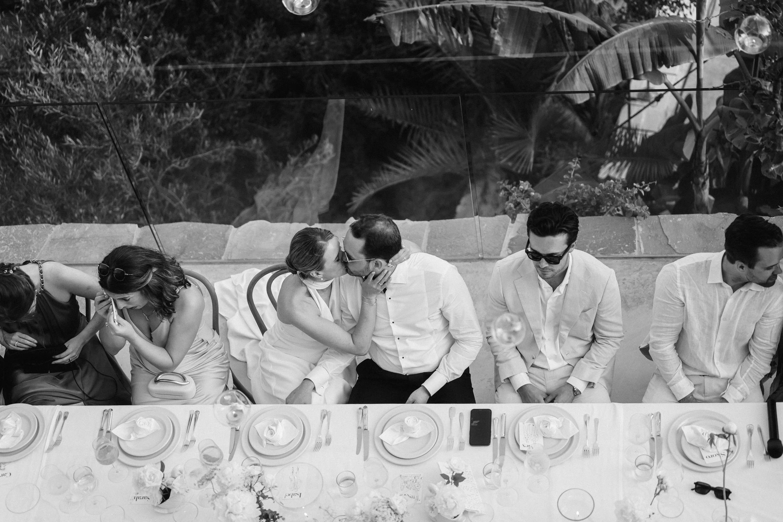 People dressed in formal attire sitting at a dining table, with a woman and man sharing a kiss. The table is set with plates, cutlery, glasses, and floral centerpieces. The photo is in black and white.