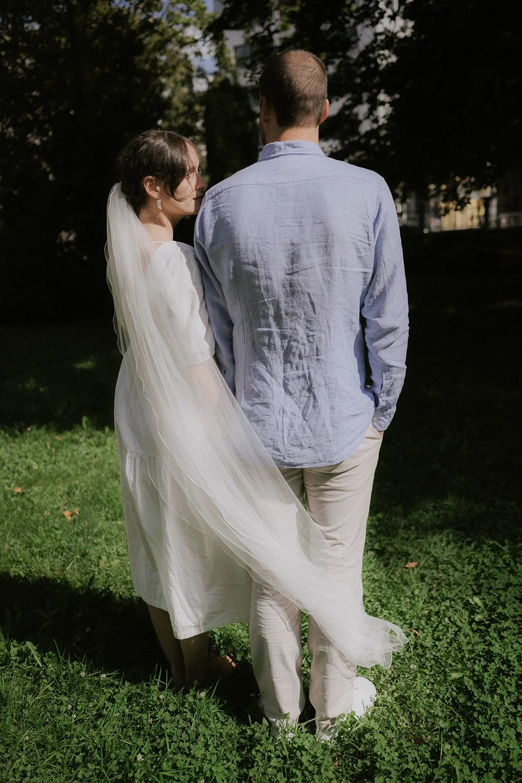 A bride and groom standing close together outdoors on a grassy area, with the bride looking at the groom, who has his back to the camera. The bride is wearing a white dress with a veil, and the groom is dressed in a light blue shirt and beige pants.