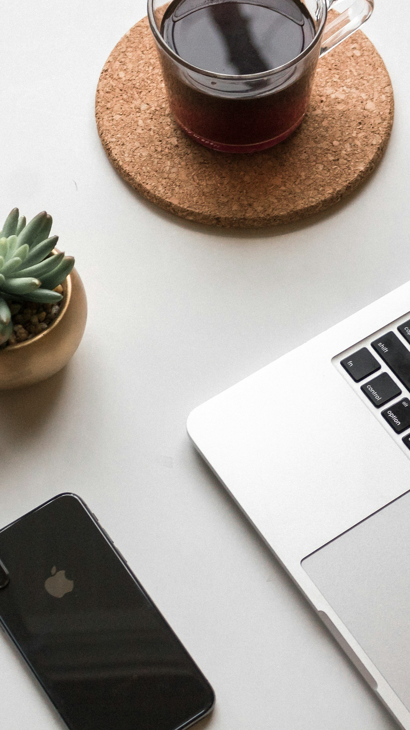 A workspace with a potted succulent plant, a cup of dark coffee on a cork coaster, part of a silver laptop, and a black iPhone on a white surface.