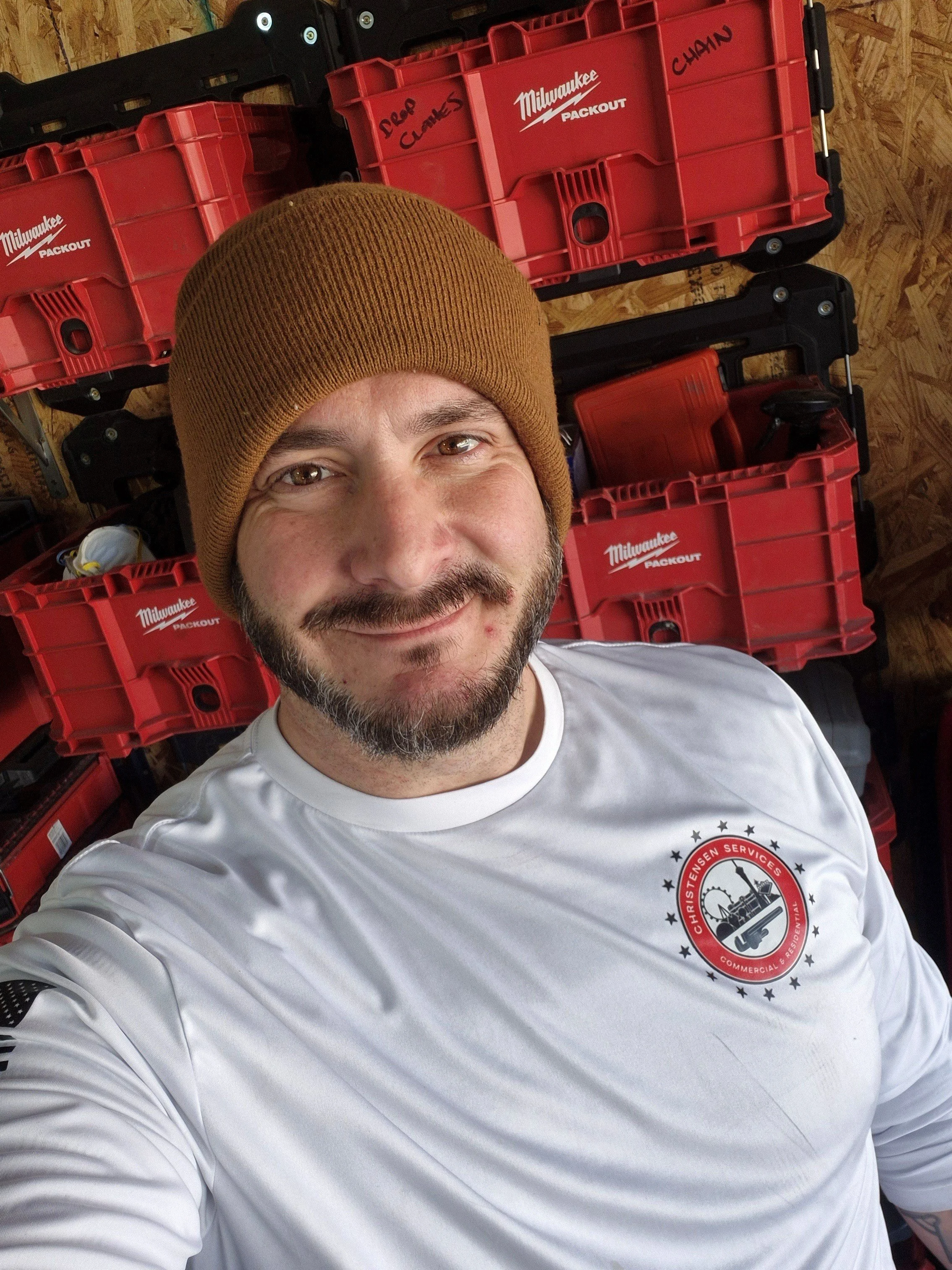 A man with a beard and short hair wearing glasses on his head and a white collared shirt, sitting indoors with a blurred background of other people and windows.