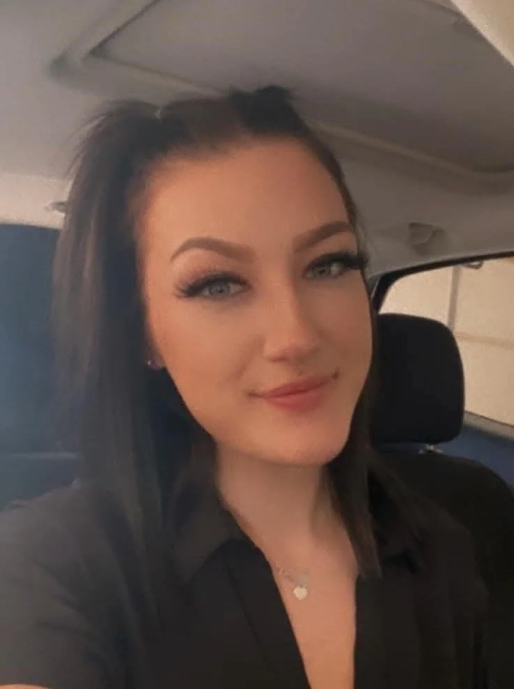A young woman with dark brown hair and blue eyes takes a selfie inside a vehicle. She is smiling slightly and has makeup on, including eyeliner and lipstick. She is wearing a black top and a necklace with a heart-shaped pendant.
