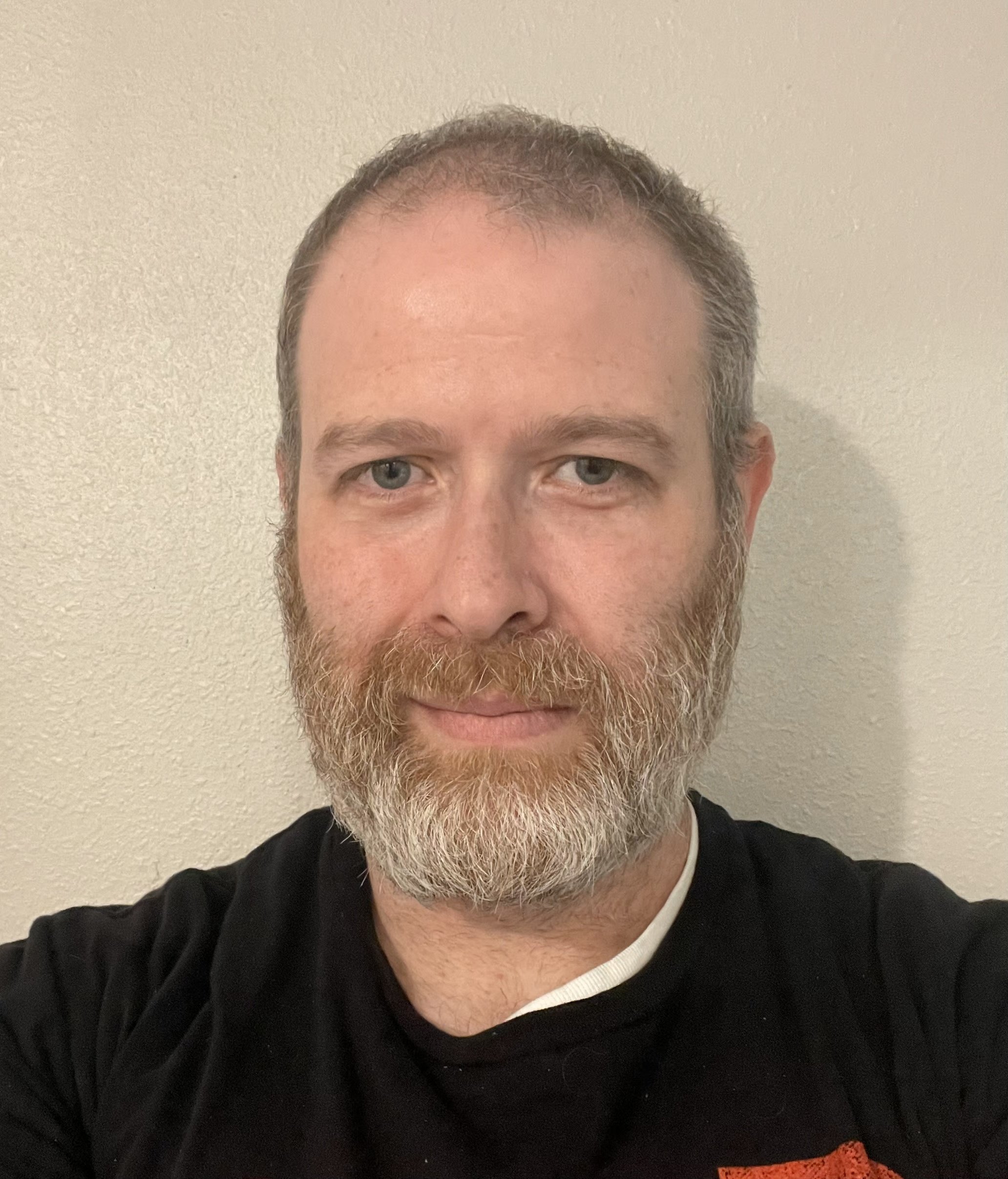 A man with a beard and short hair taking a selfie against a plain wall.