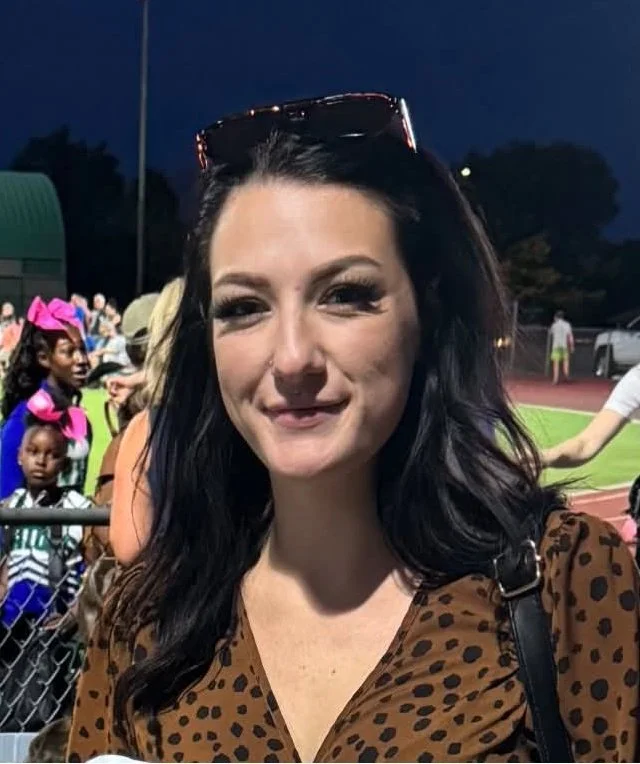 A young woman with dark brown hair and blue eyes takes a selfie inside a vehicle. She is smiling slightly and has makeup on, including eyeliner and lipstick. She is wearing a black top and a necklace with a heart-shaped pendant.