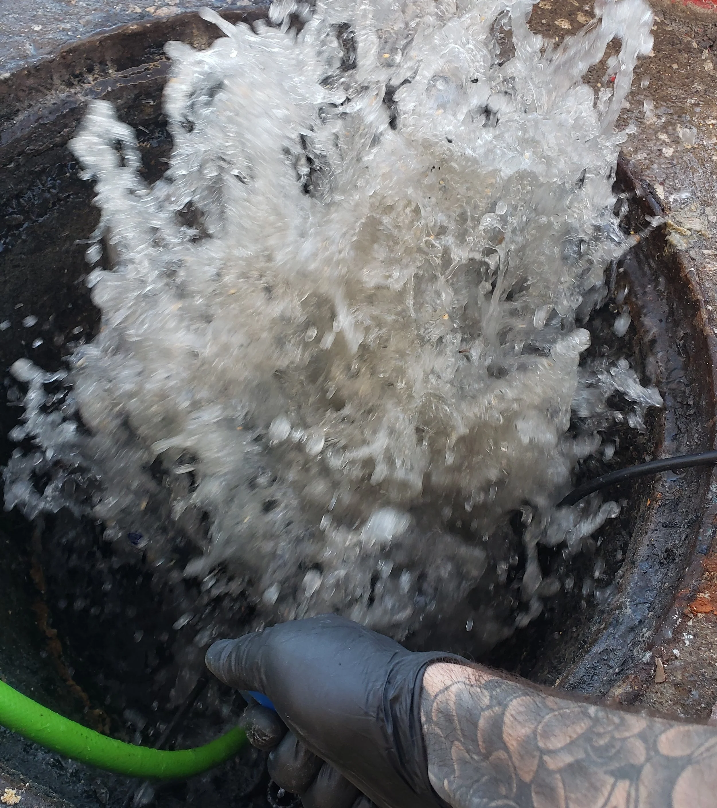 A person wearing a black glove with a tattooed wrist is holding a green hose that is spraying water into an open manhole, causing water to splash out.