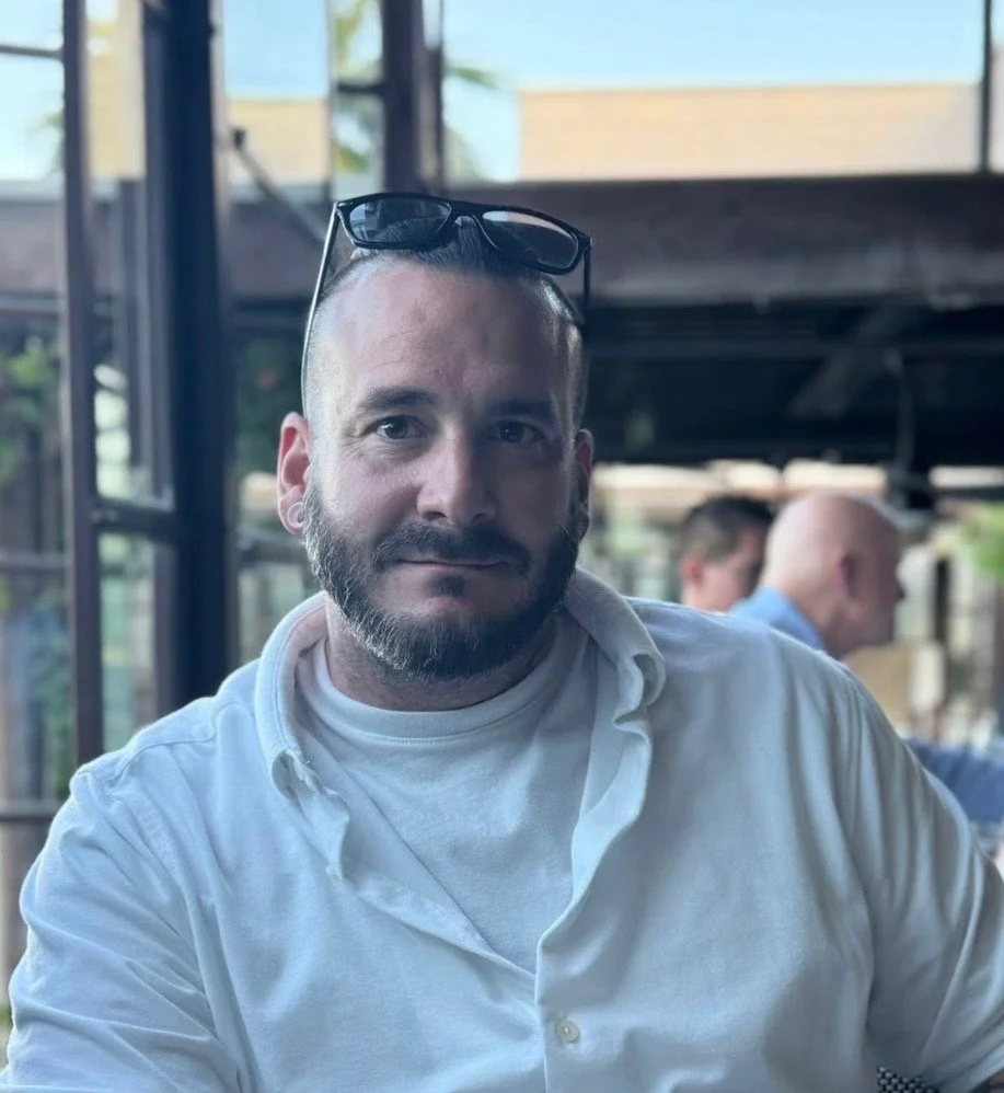 A man with a beard and short hair wearing glasses on his head and a white collared shirt, sitting indoors with a blurred background of other people and windows.