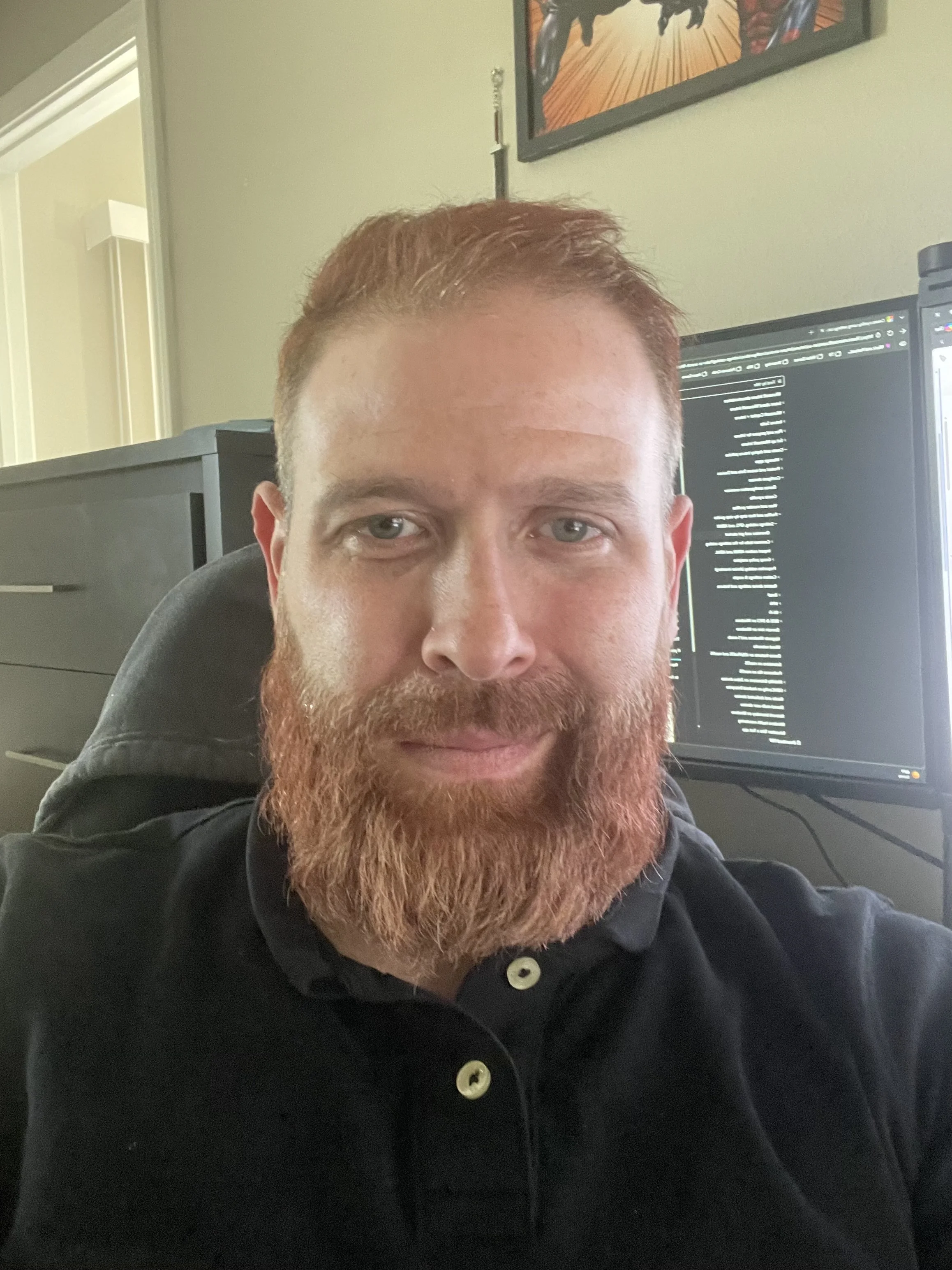 A man with a beard and short hair taking a selfie against a plain wall.