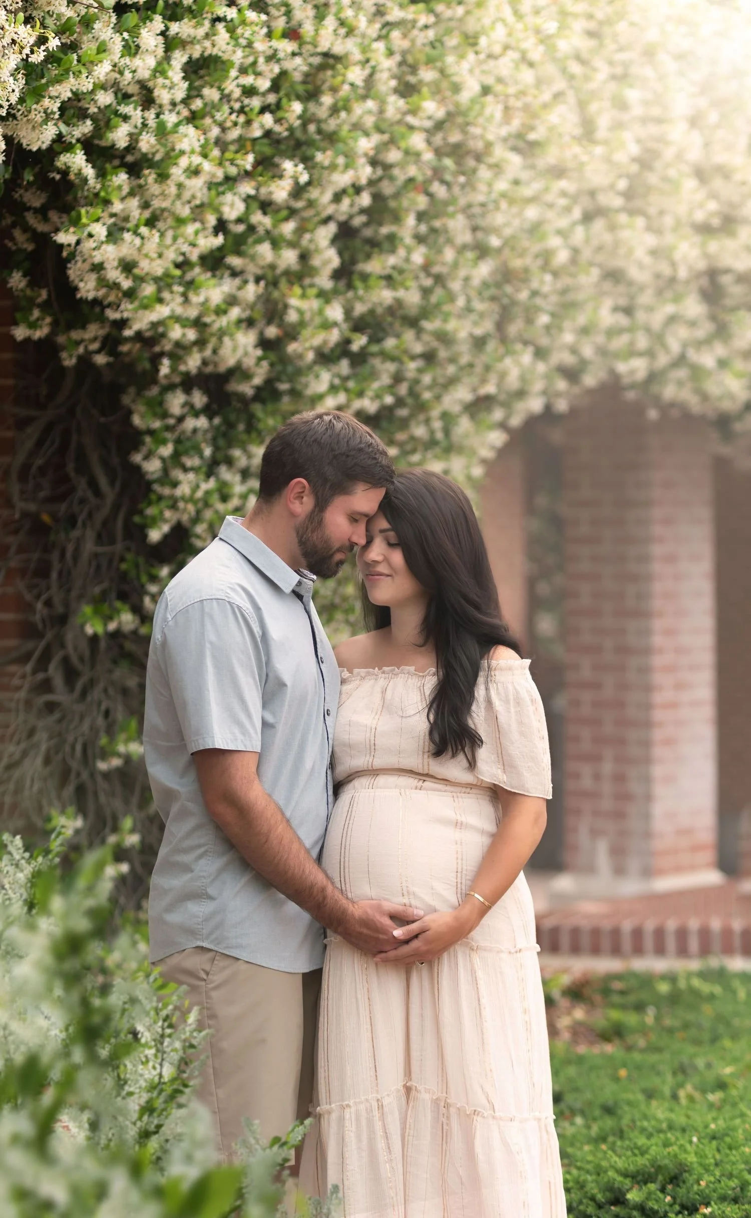 A pregnant woman and a man standing close together outdoors, holding hands over her pregnant belly, with eyes closed and heads touching, near a flowering bush and brick structure.