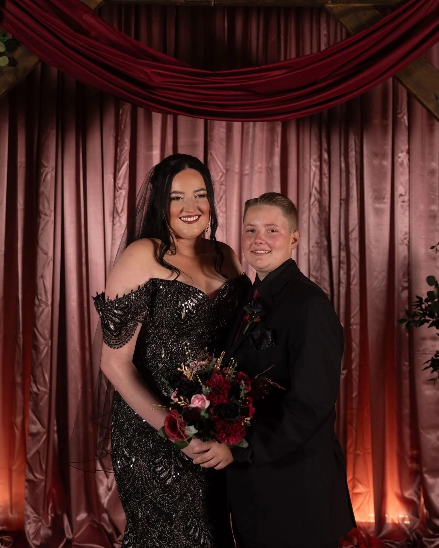 A beautiful photo taken by Trisha Disney-Linder of a lesbian couple. A woman in a black, sequined, off-the-shoulder evening gown holding a bouquet of pink flowers poses with her wife in a black tuxedo against a backdrop of pink and red curtains.