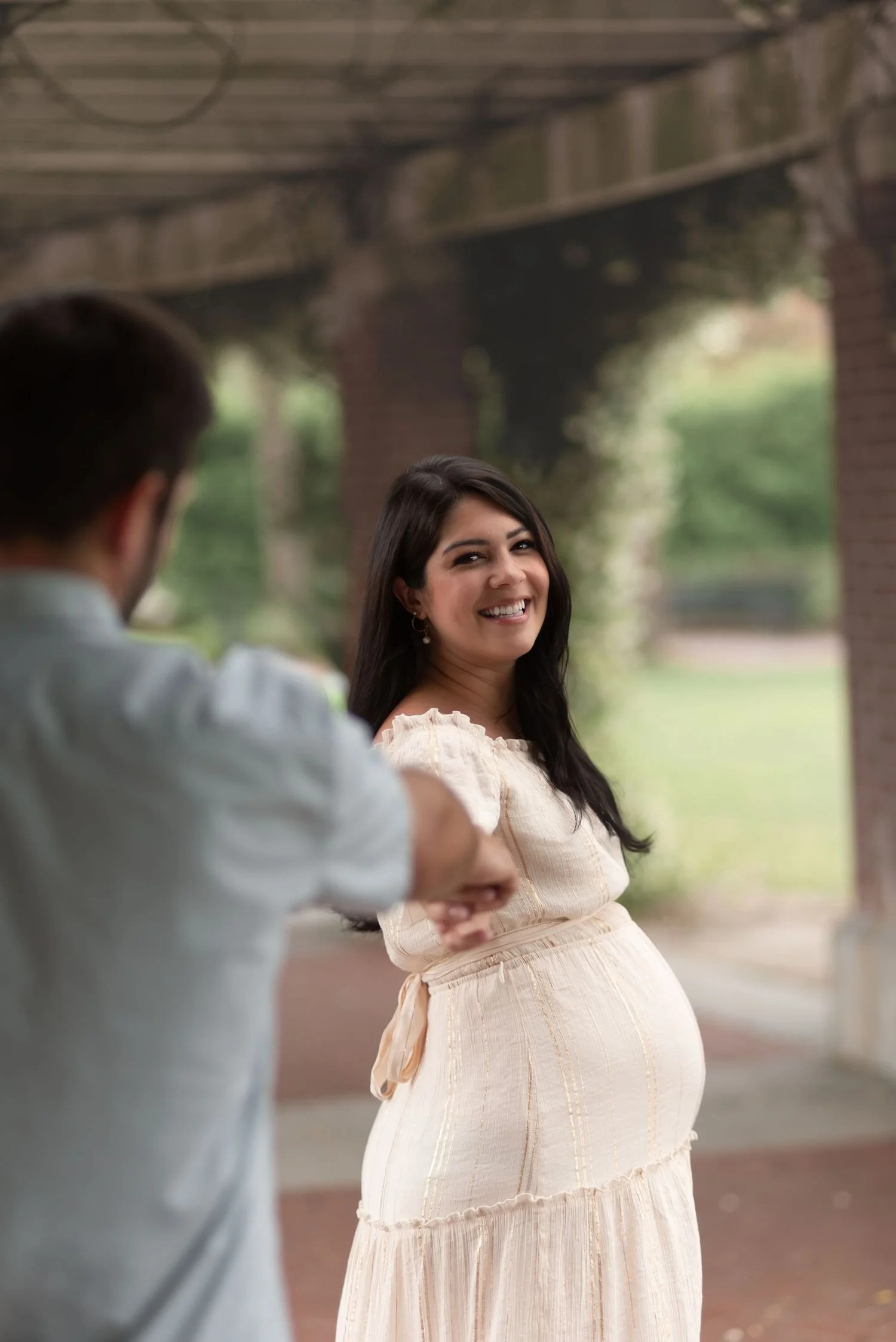 A pregnant woman smiling and looking at the camera while standing outdoors under a bridge, with a man in the foreground pointing at her.